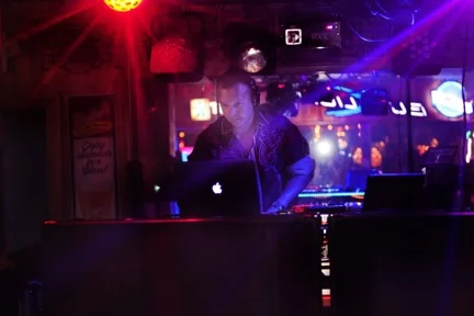DJ working behind a laptop and mixer under red and blue lights.