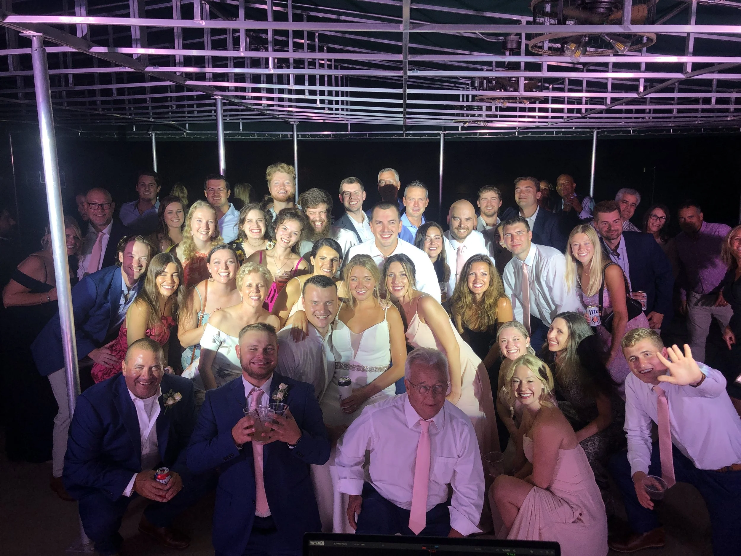 Wedding guests gathered for a group photo beneath purple-lit metal ceiling beams.