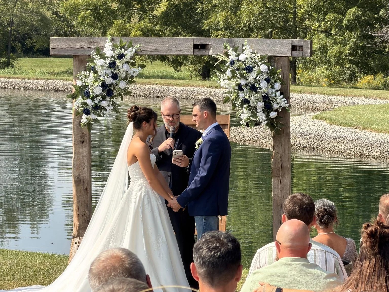 Couple holding hands with an officiant beneath a flower-decorated wooden arch beside the water.