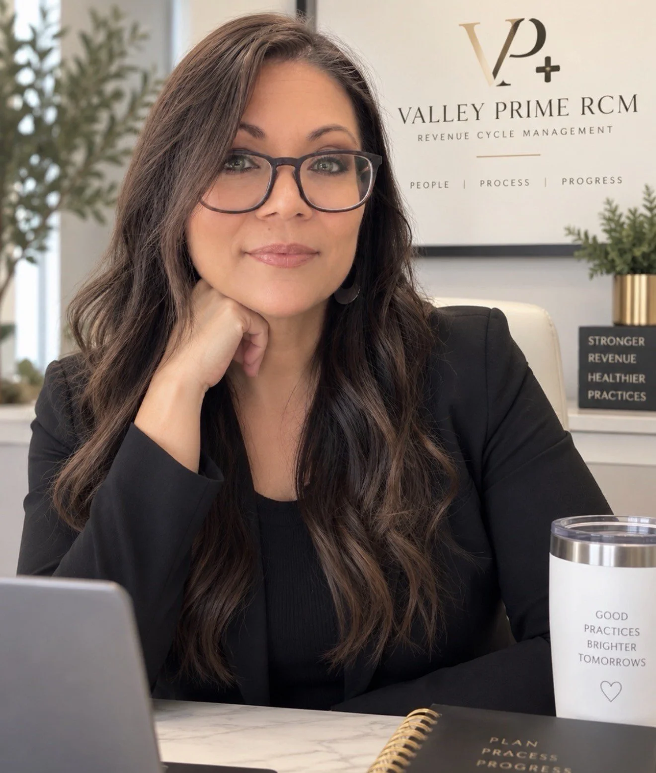 A woman with long brown hair, glasses, and a black blazer sits at a desk with her chin resting on her hand. In the background, a sign reads "Valley Prime RCM" and an inspirational quote on a white tumbler says "Good practices brighter tomorrows."