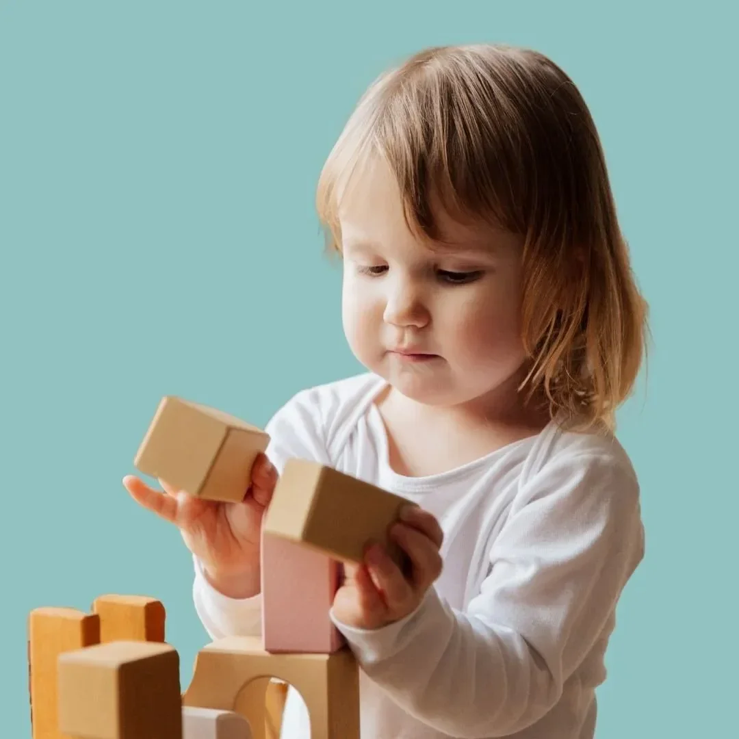 A young child with brown hair wearing a white long-sleeve shirt playing with wooden building blocks against a light blue background.