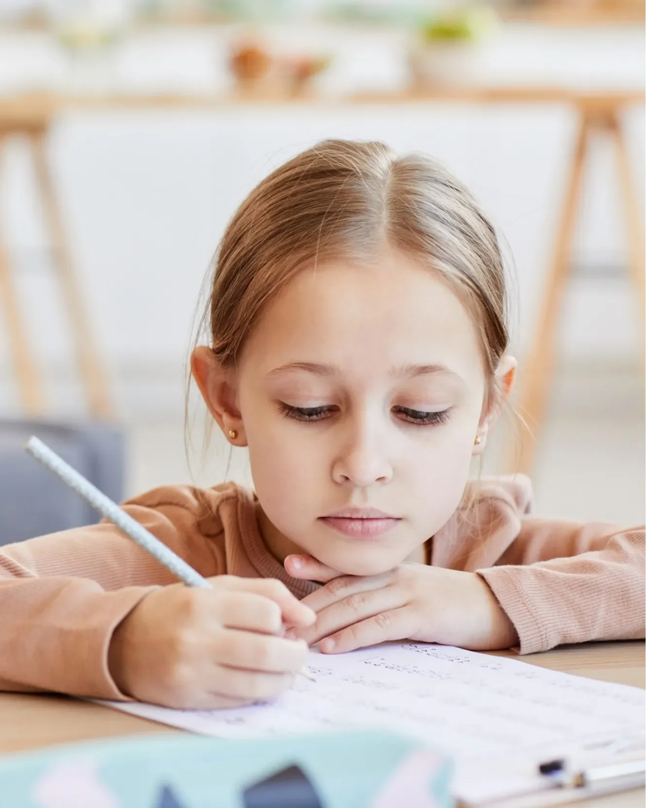 A young girl with light skin and light brown hair, wearing a peach-colored long-sleeve shirt, is writing on a piece of paper at a desk in a well-lit room, with her head resting on her crossed arms.