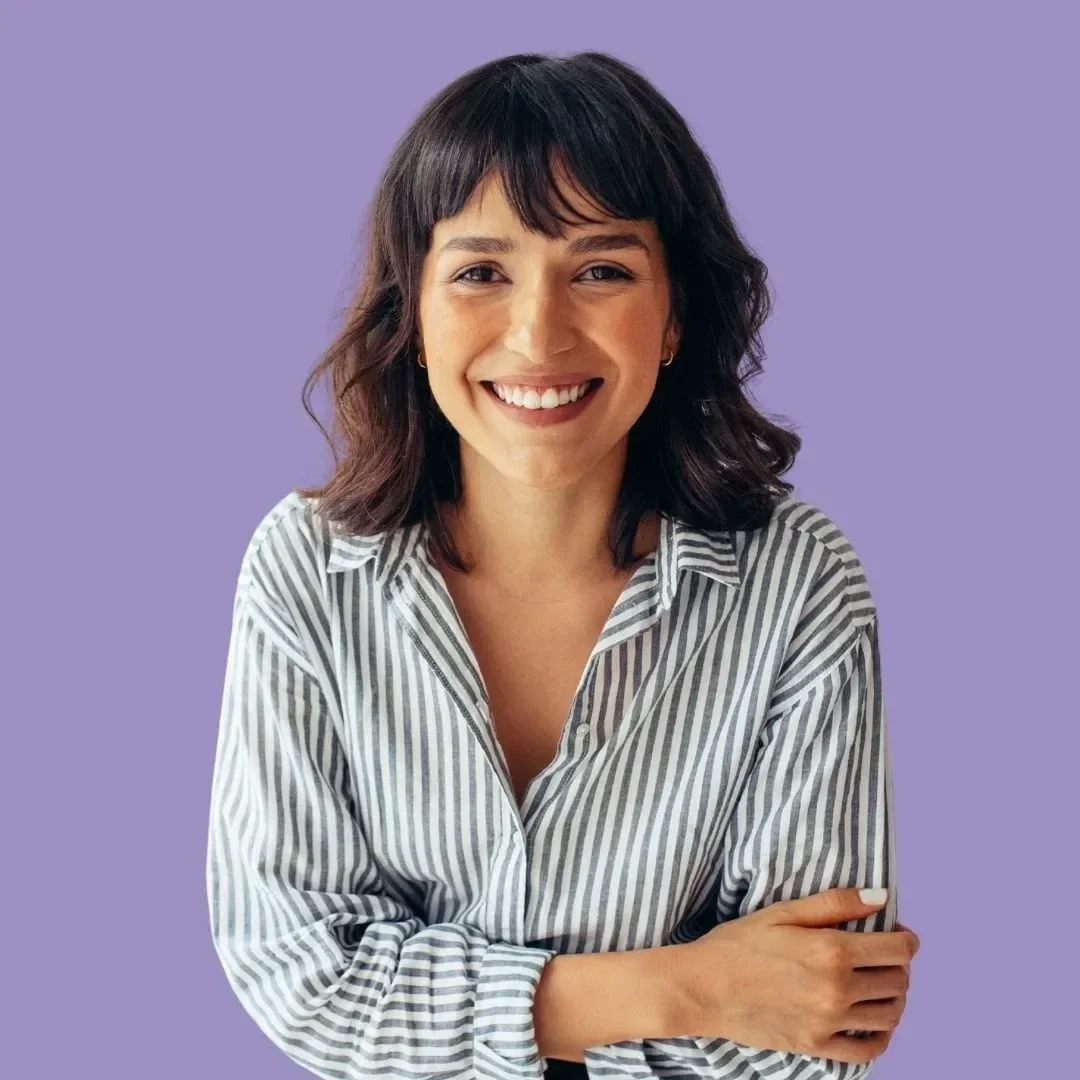 A woman with dark brown, wavy hair and bangs, smiling, wearing a white and grey striped shirt against a lavender background.