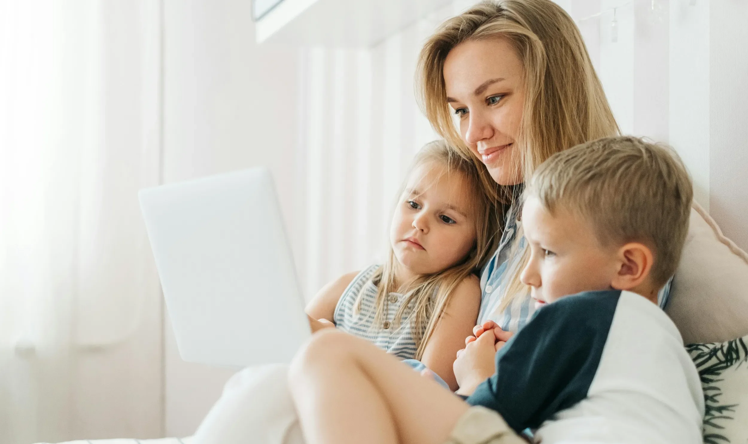 A woman with long hair sitting on a couch with two young children, a girl and a boy, in a bright, modern room, possibly watching a digital device.