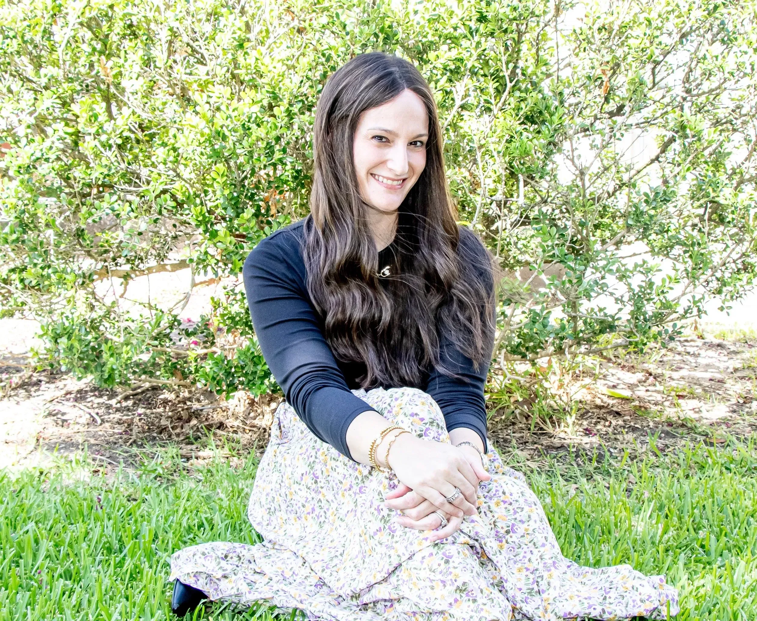 Dr. Erika Westreich with long dark hair sitting cross-legged on grass in front of green shrubbery, smiling at camera, wearing a black top and a floral skirt, accessorized with jewelry.