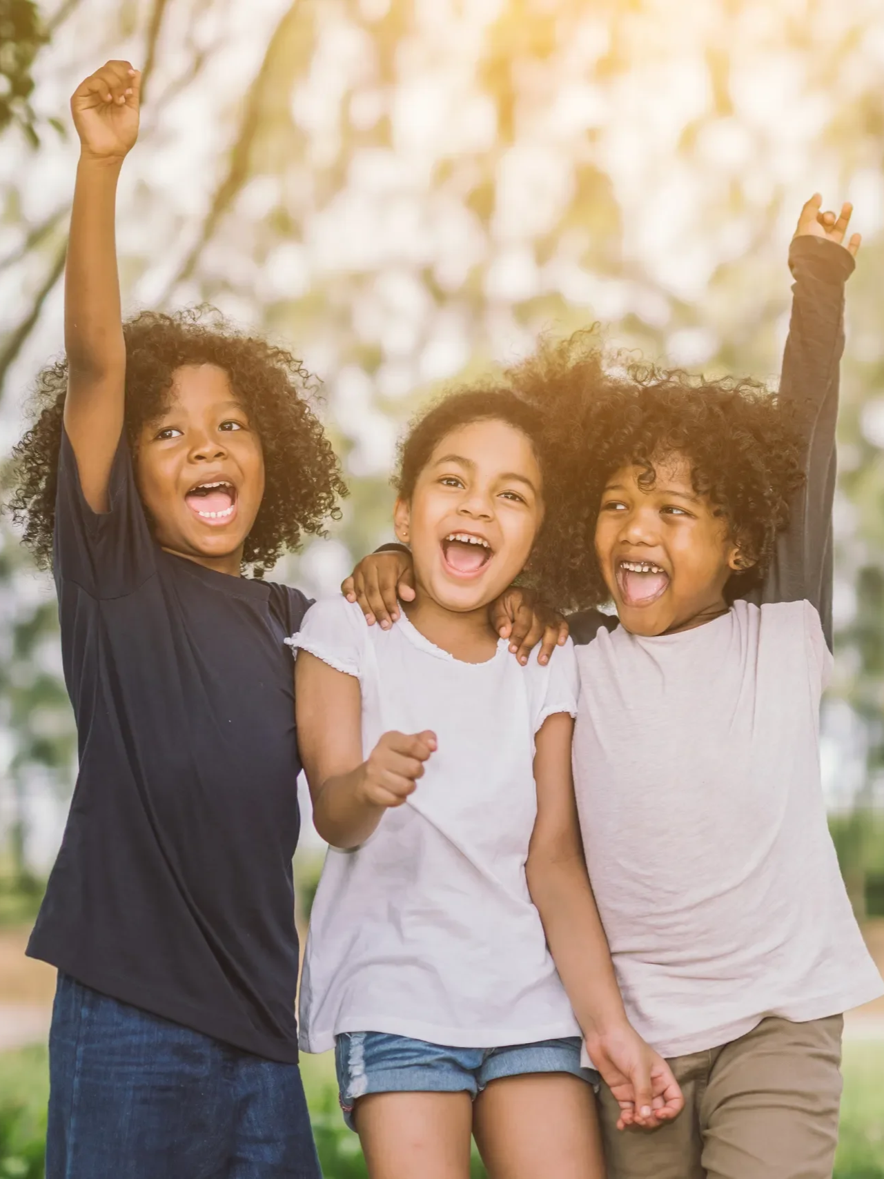 Three children, two girls and a boy, happily embracing outdoors with cheerful expressions, sunlight filtering through trees in the background.