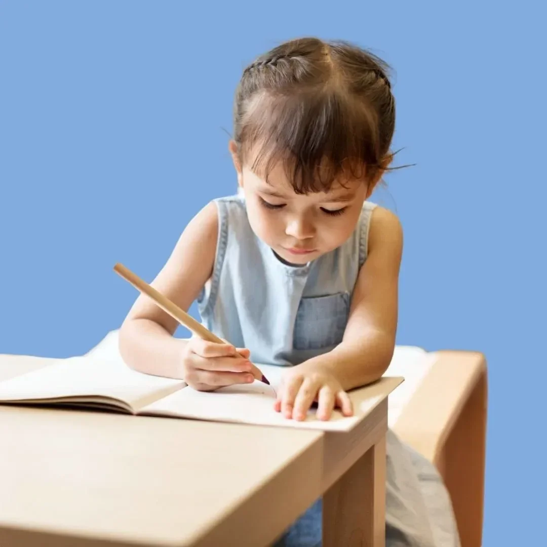 A young girl with brown hair and bangs, wearing a light blue sleeveless top, is sitting at a wooden table and drawing or writing in a notebook with a colored pencil against a plain blue background.