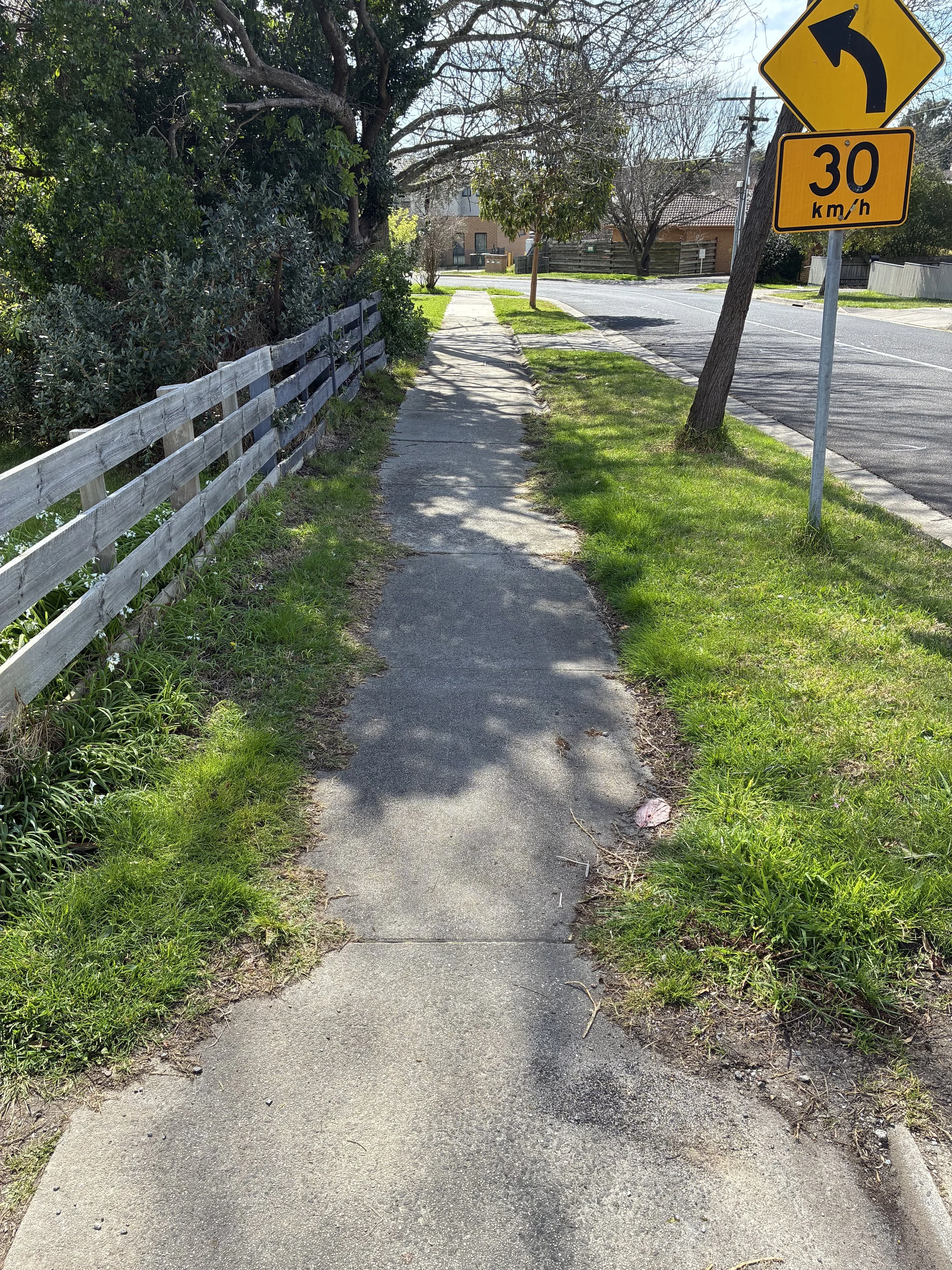 Sidewalk with a wooden fence on the left, grass and trees on the right, and street signs indicating a left turn and a speed limit of 30 km/h. Residential buildings and utility poles are visible in the background.