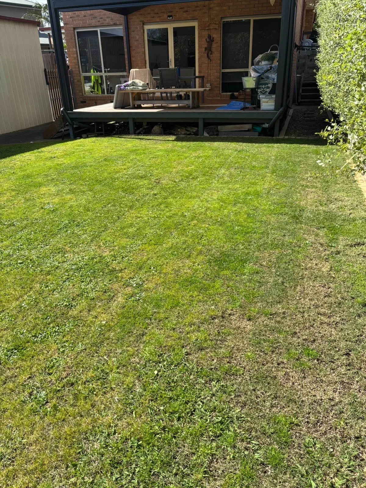A backyard with a green lawn and a raised wooden deck attached to a brick house, with a sliding glass door and a window. The deck has outdoor furniture, some items covered with a towel, and various miscellaneous objects. A staircase on the right lead