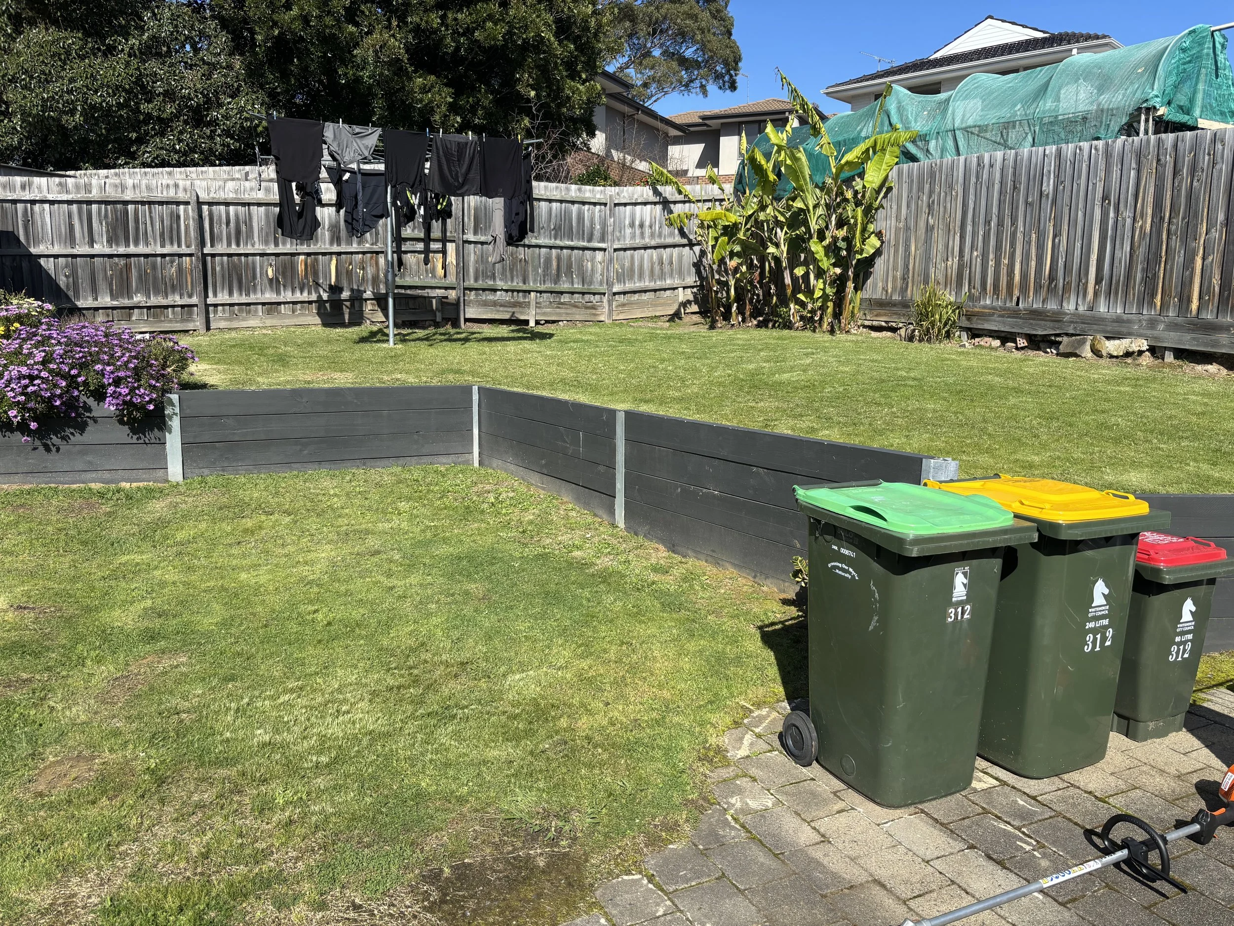 A backyard patio with a grassy lawn, laundry hanging on a clothesline, green and yellow trash bins, a wooden fence, flowers, and outdoor furniture.