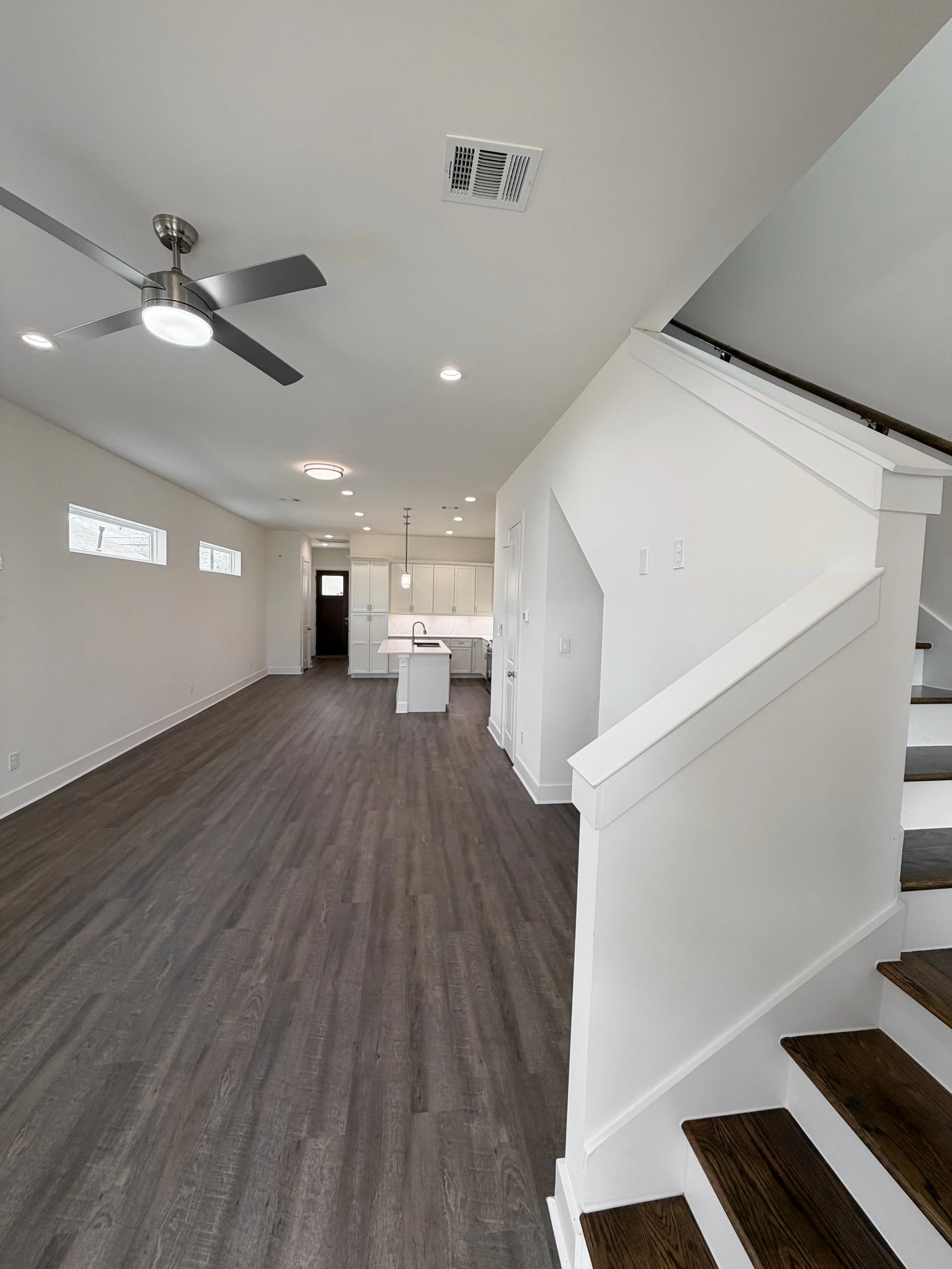 Interior view of a modern, open-plan living area with white walls, dark wood flooring, a ceiling fan, reccessed lighting, and a staircase leading upstairs.