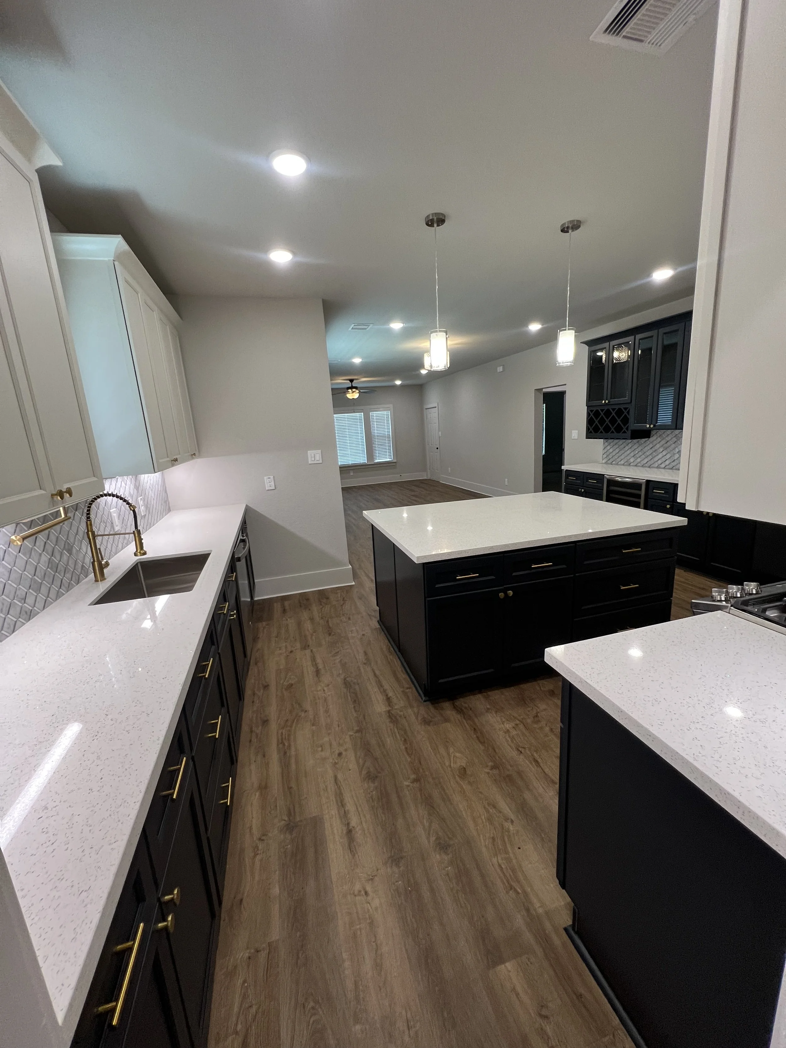 Inside view of a modern kitchen with white upper cabinets, black lower cabinets, white countertops, gold handles, a stainless steel sink, and wooden flooring. There are pendant lights hanging over the island, with recessed lighting in the ceiling, and a view into a dining or living area in the background.