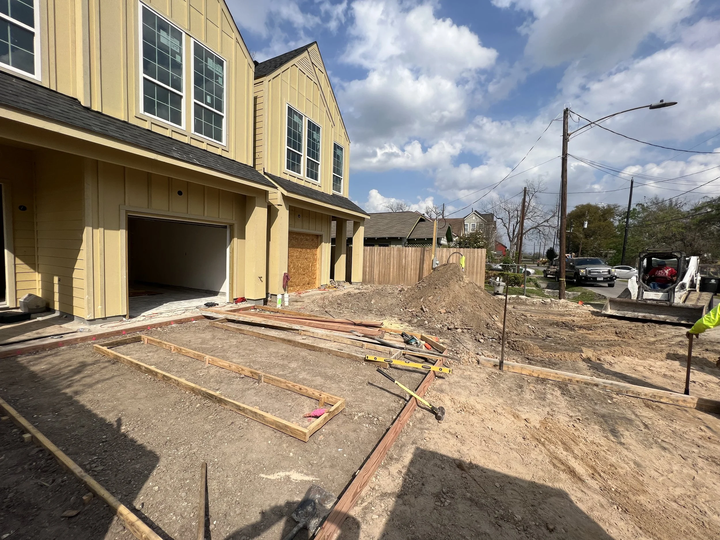 A residential construction site with framing laid out for a concrete driveway, yellow house with large windows in the background, and a partly cloudy sky.