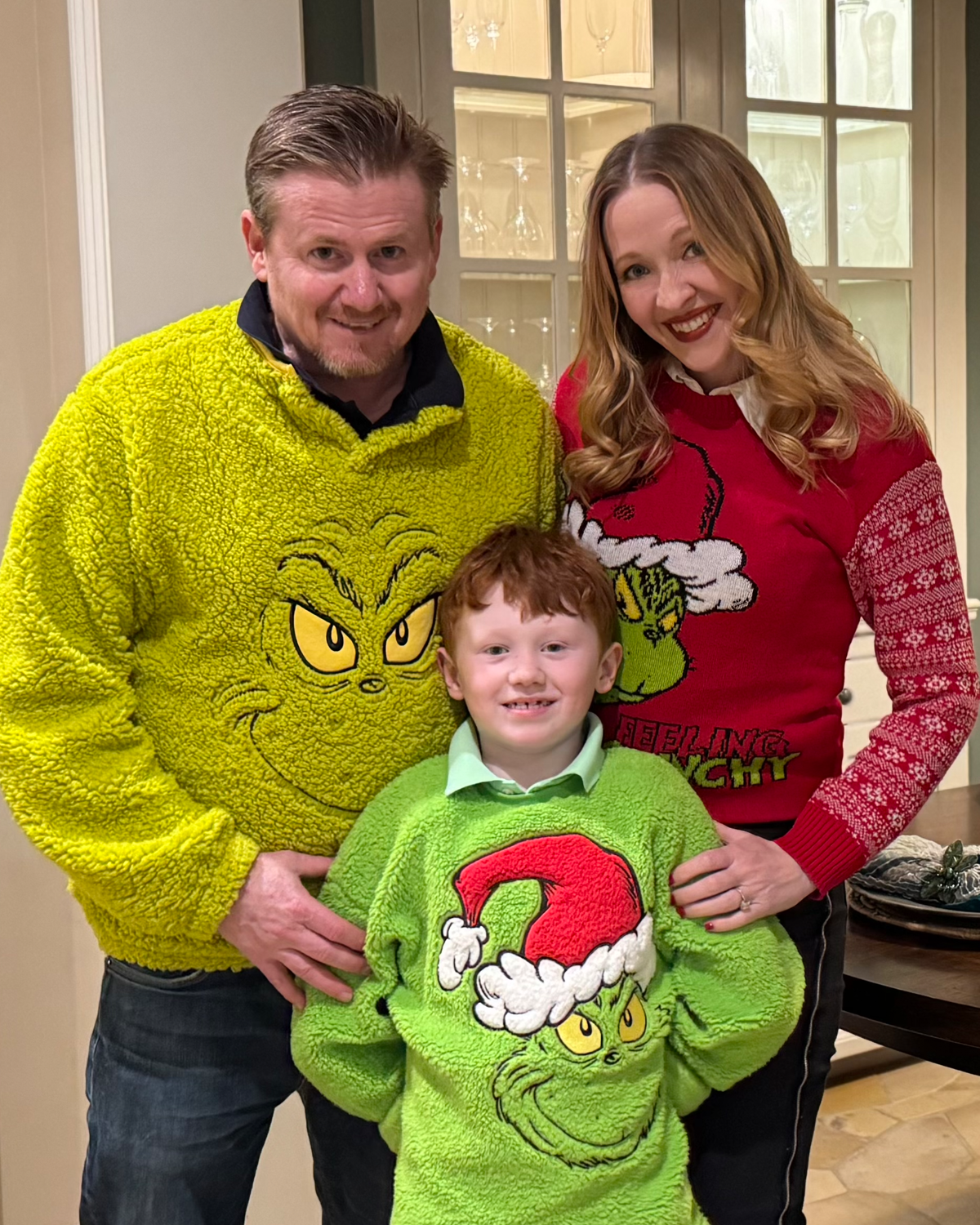 A smiling family of three dressed in Grinch-themed sweaters in a cozy indoor setting.