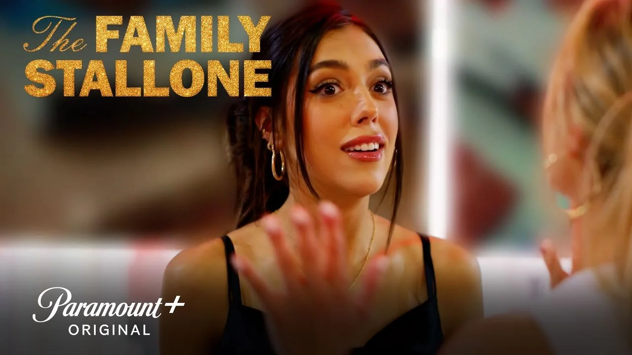 A woman with dark hair and hoop earrings engaging in a conversation, with the words 'The Family Stallone' written in gold text in the upper left corner and the Paramount+ logo in the lower left corner.