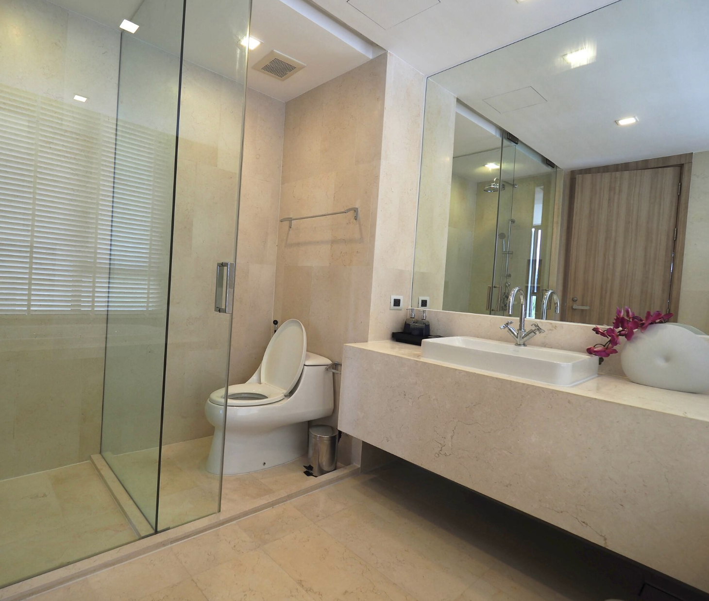Modern bathroom with glass-enclosed shower, toilet with open lid, large mirror, beige countertops, and a decorative flower arrangement in a white vase.