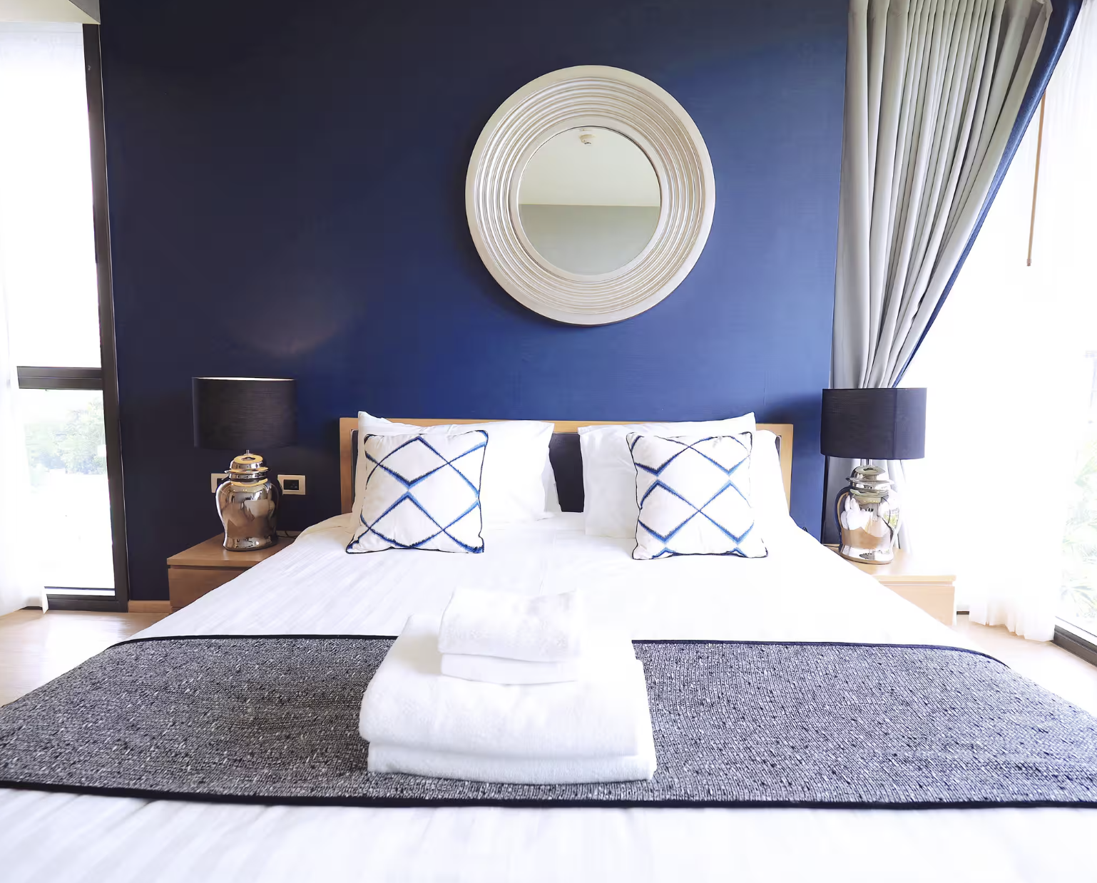 A modern bedroom with a white bed, dark blue wall, round decorative mirror, two black table lamps, and white curtains.