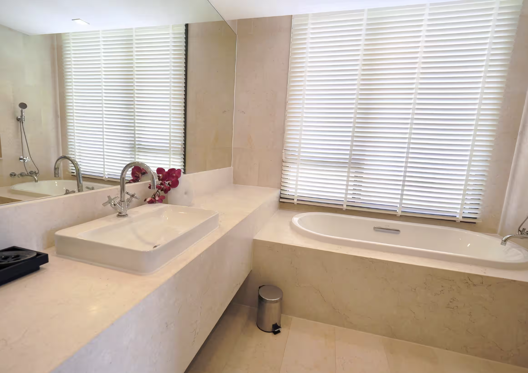 Modern bathroom with beige countertops, a white rectangular sink with a chrome faucet, a large mirror, pink flowers, a bathtub, and large windows with white blinds.