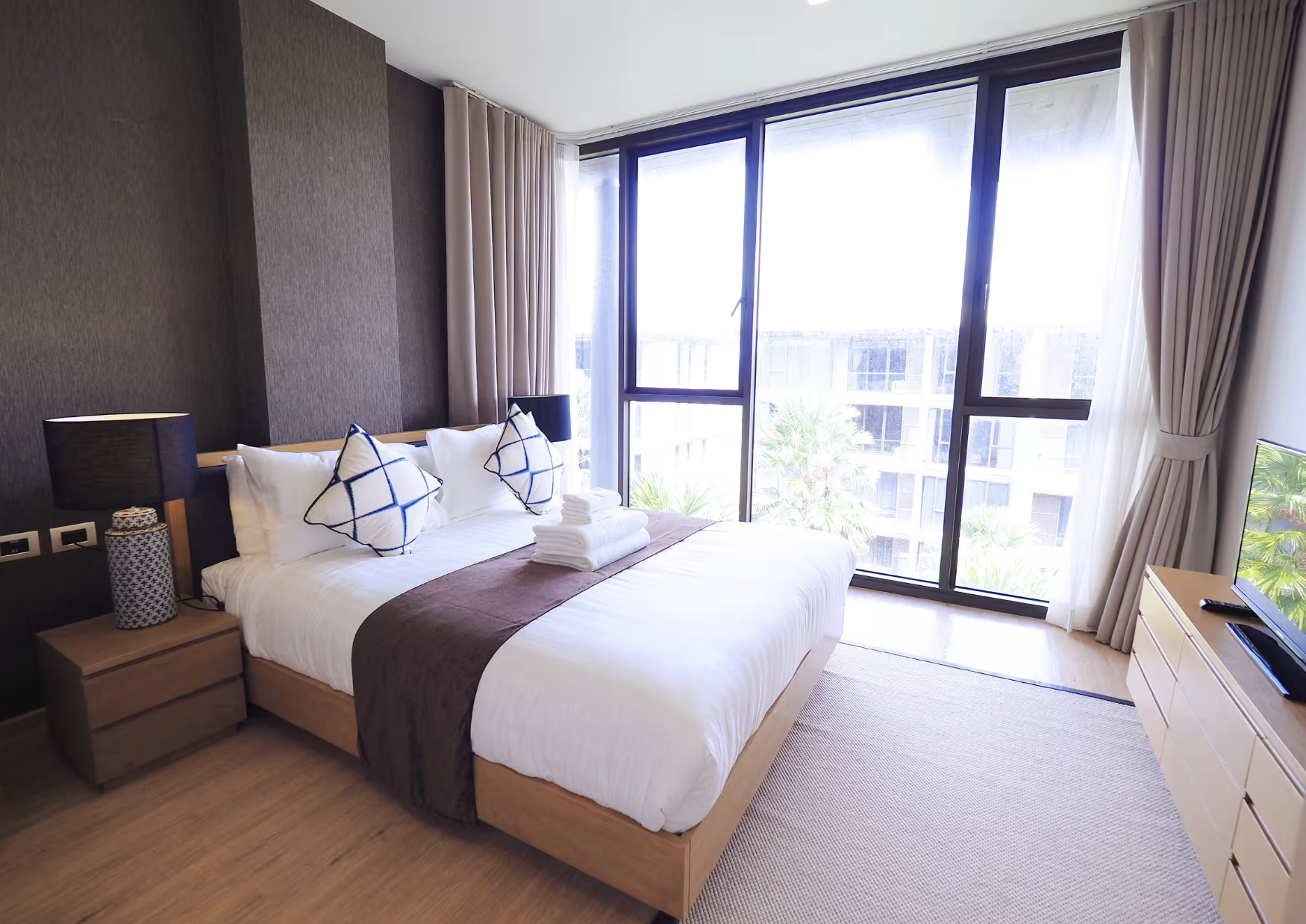 Modern hotel room with large floor-to-ceiling windows showing a view of palm trees outside. The bed has white linens, decorative pillows with blue and white designs, a brown throw blanket, and neatly folded towels on top. There are bedside lamps on w