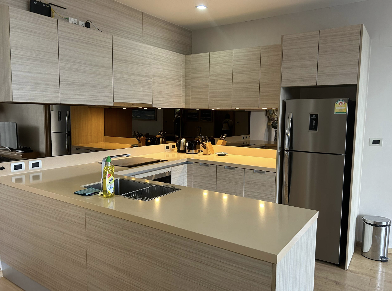 Modern kitchen with light wood cabinets, a beige countertop, stainless steel refrigerator, and various small appliances on the counter including a toaster and electric kettle. A bottle of dish soap and a sponge are near the sink.