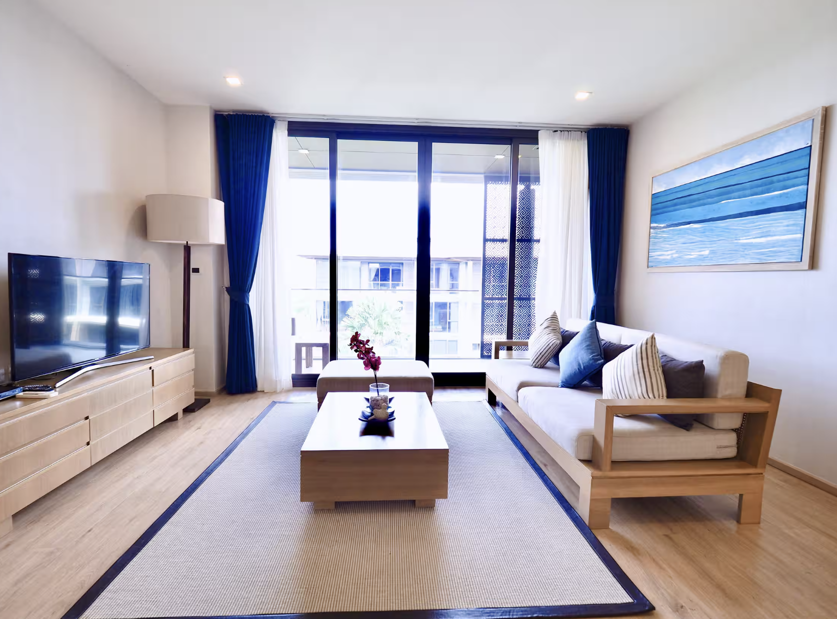 Living room with a beige sofa decorated with striped and blue pillows, a wooden coffee table with a flower vase, a large window with white and blue curtains, a flat-screen TV on a wooden cabinet, a tall floor lamp, and a painting of the sea on the wa