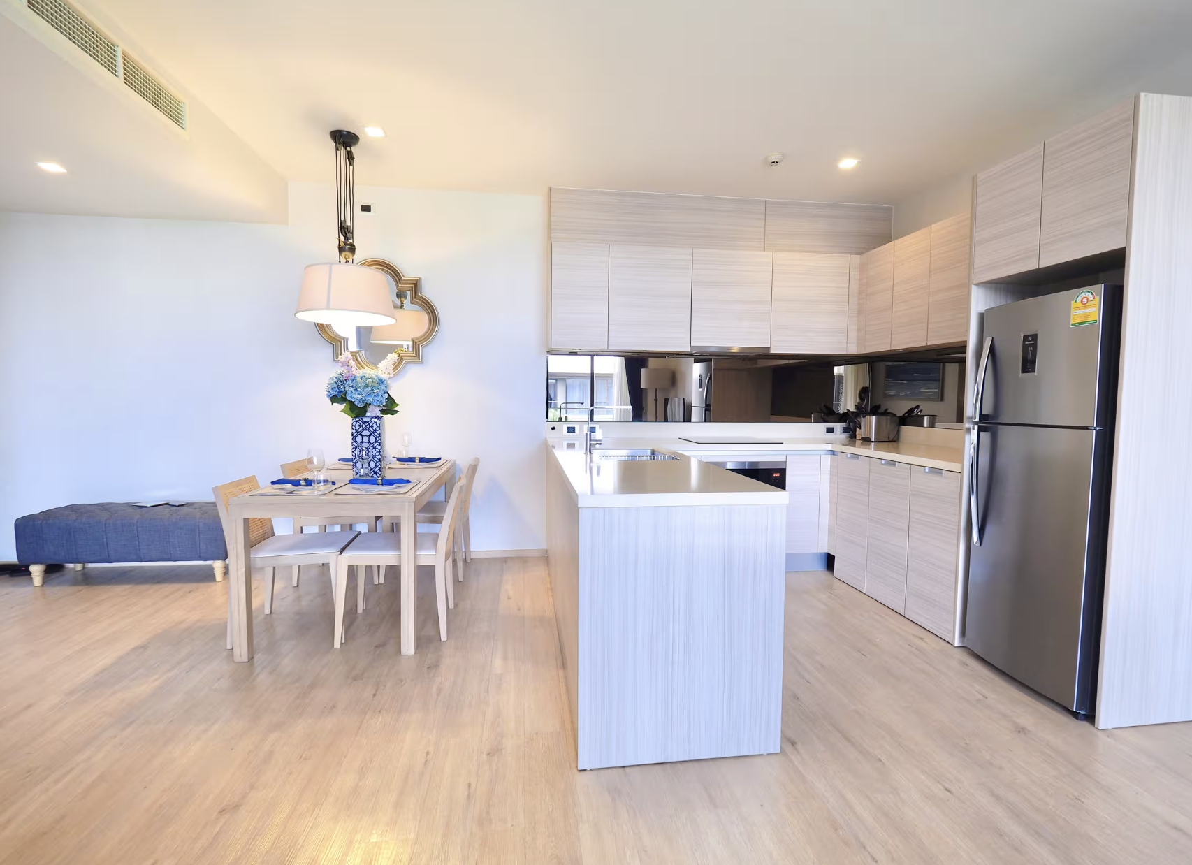 Modern kitchen with light wood cabinets, stainless steel refrigerator, white countertop, and a dining area with a wooden table, four chairs, and a vase with blue hydrangeas. The space is brightly lit with ceiling lights.