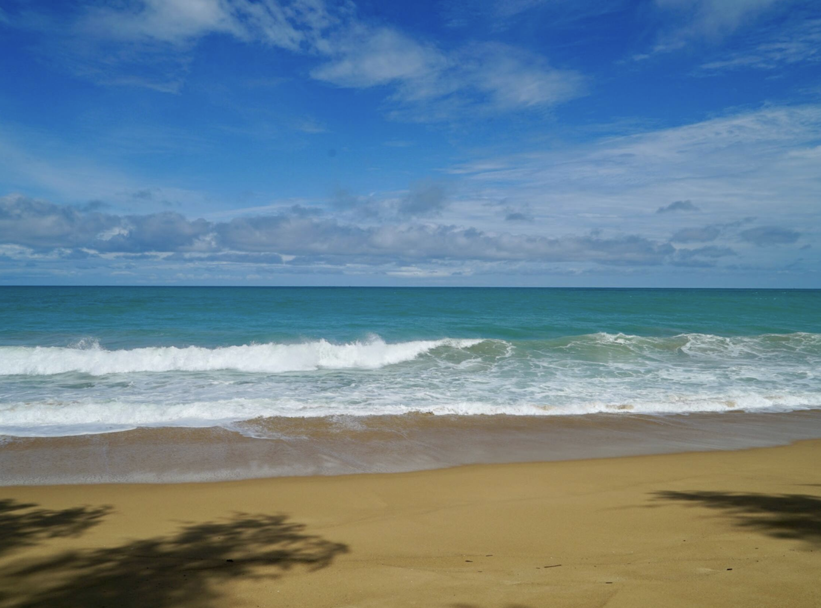 A sandy beach with ocean waves and a partly cloudy blue sky.