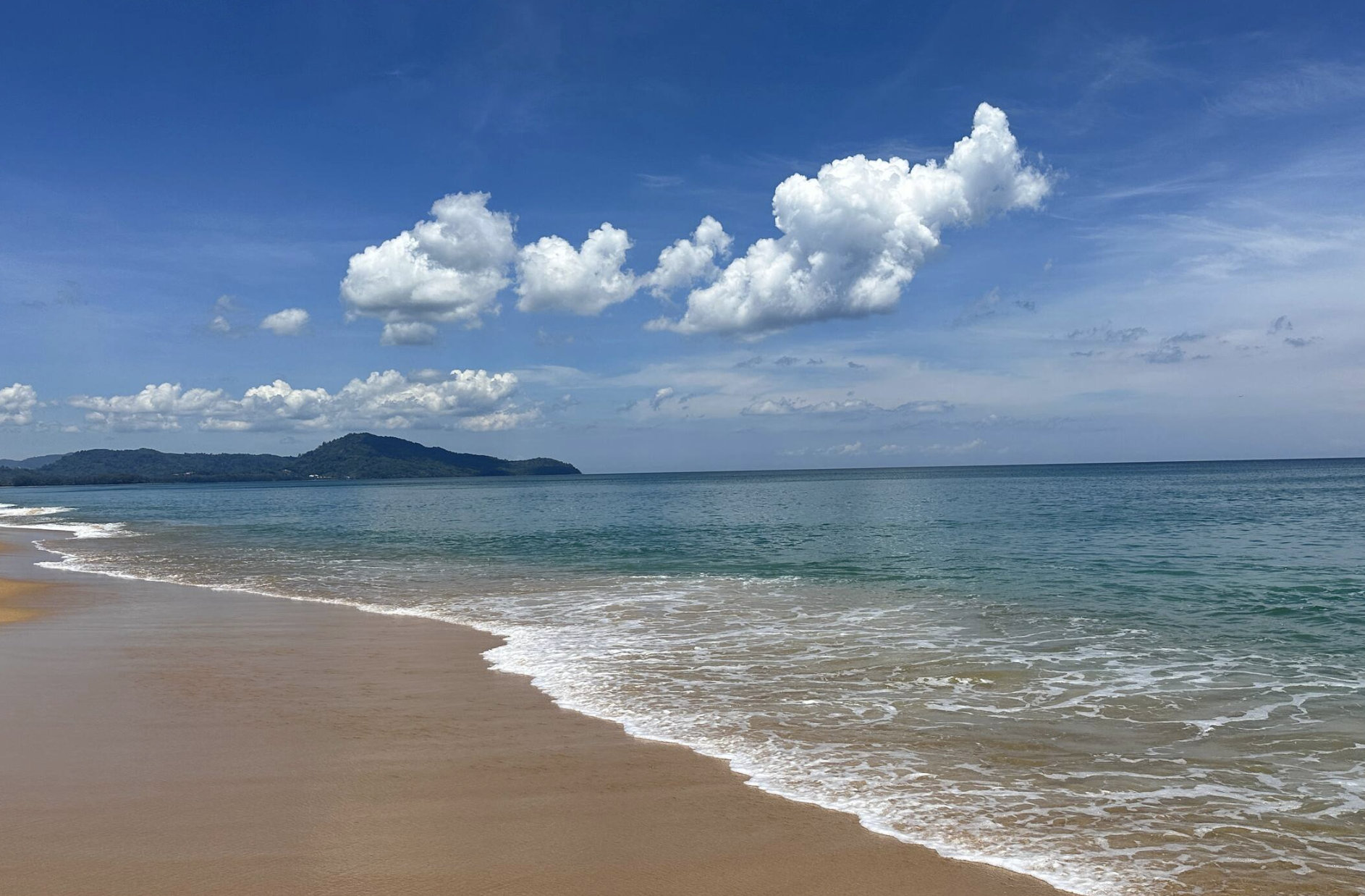 A sandy beach with gentle waves along the shore, a calm ocean, a distant green hill, and a partly cloudy blue sky.