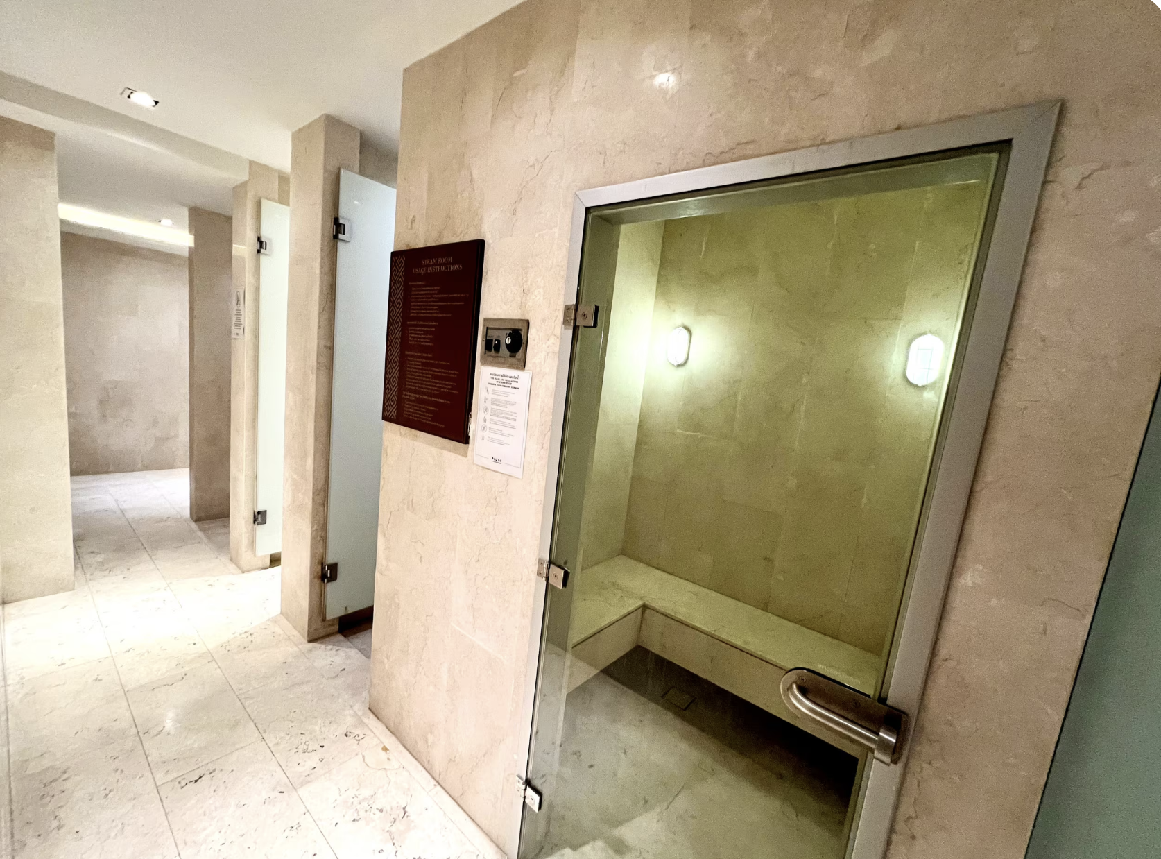 A glass enclosed steam room with a beige interior and a rounded corner. There is a light on the wall and a bench inside. Nearby, a fire alarm pull station and a red information sign are mounted on a marble wall. The surrounding area features beige ma
