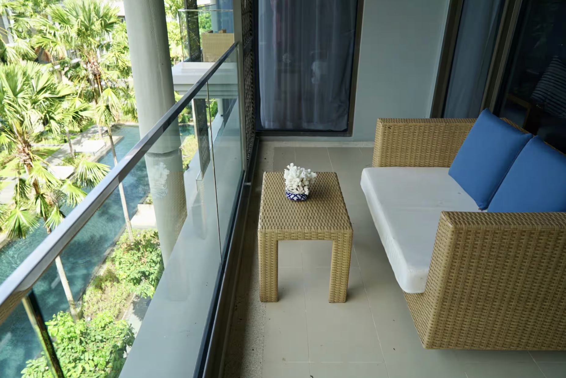 Balcony with wicker chair, small table with white coral decoration, blue cushion, view of lush green trees and swimming pool outside.