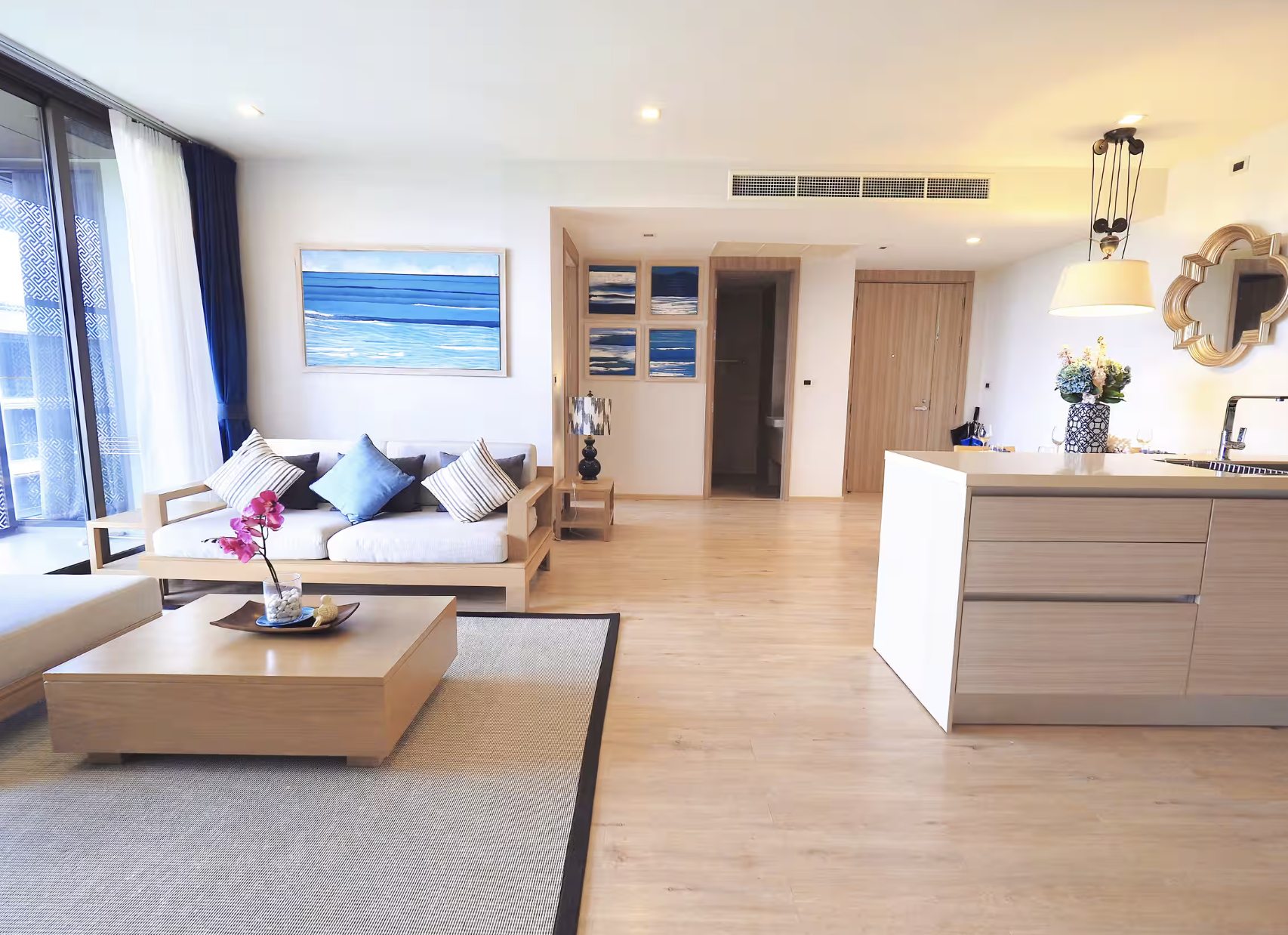 Living room with a beige couch, decorative pillows, a wooden coffee table with a pink orchid, a painting with blue ocean views on the wall, and large glass sliding doors with white and blue curtains leading to a balcony.