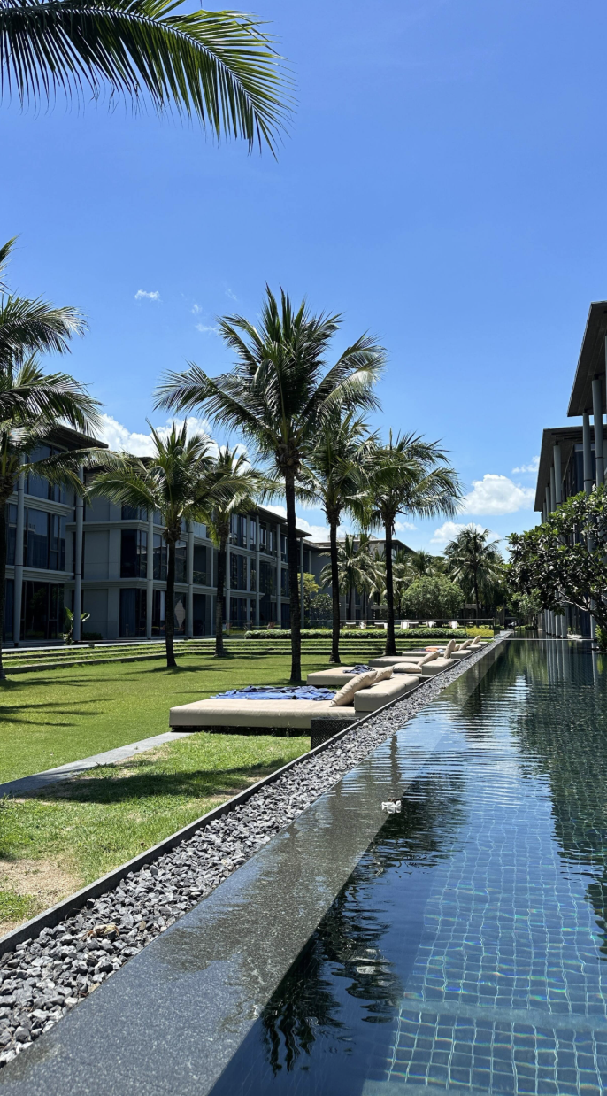 Resort pool area with lounge chairs, palm trees, modern buildings, and a clear blue sky.