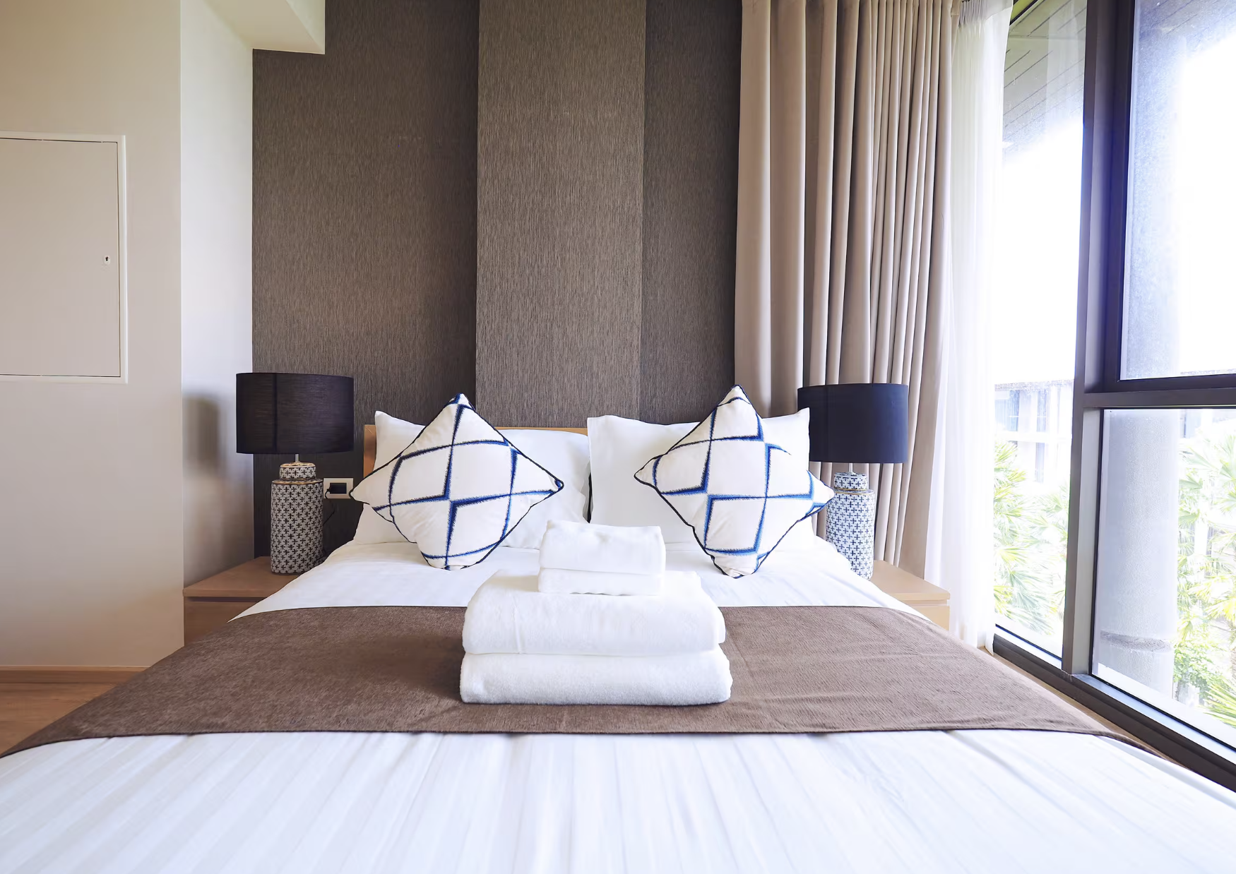 A neatly made bed in a modern hotel room with white sheets, brown blanket, and three decorative pillows with blue and white patterns. Two black bedside lamps sit on nightstands beside the bed. A window with beige curtains and a sliding glass door are visible, revealing greenery outside.