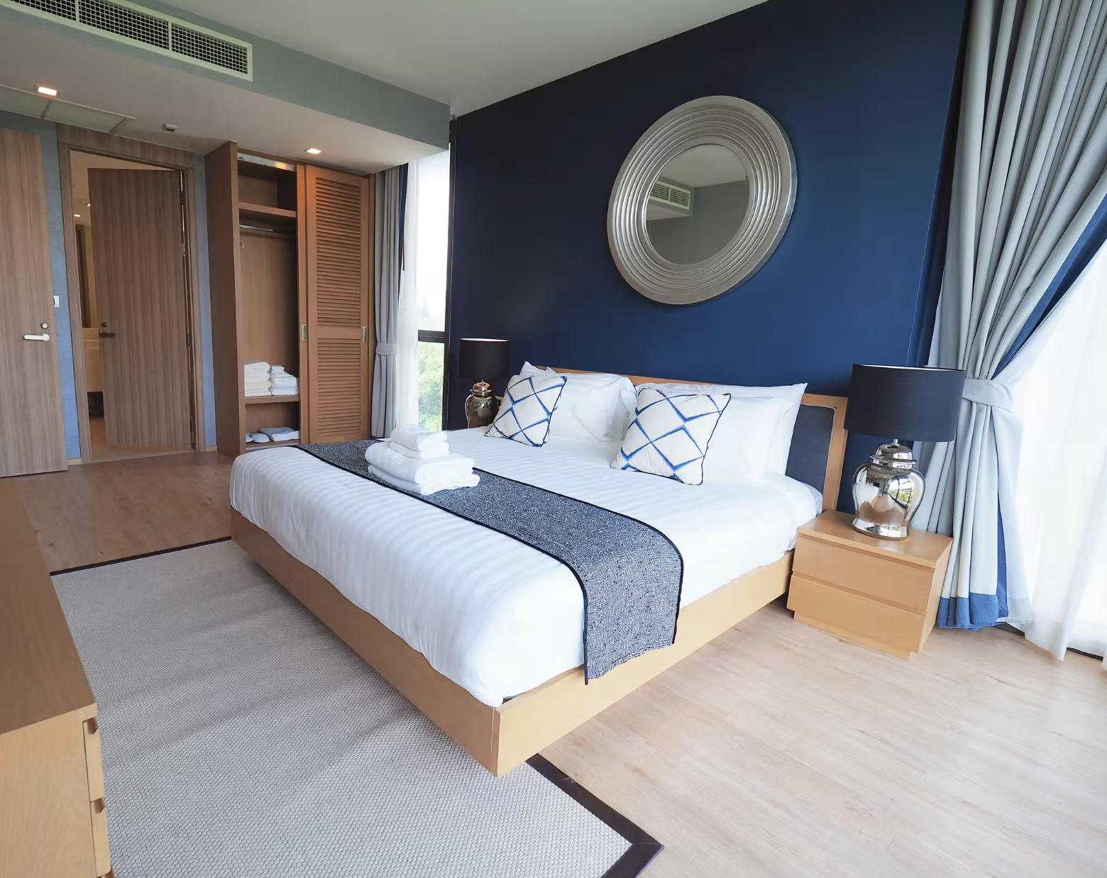 Modern hotel room with a neatly made bed, blue accent wall, round mirror, bedside tables with black lamps, and a window with curtains.