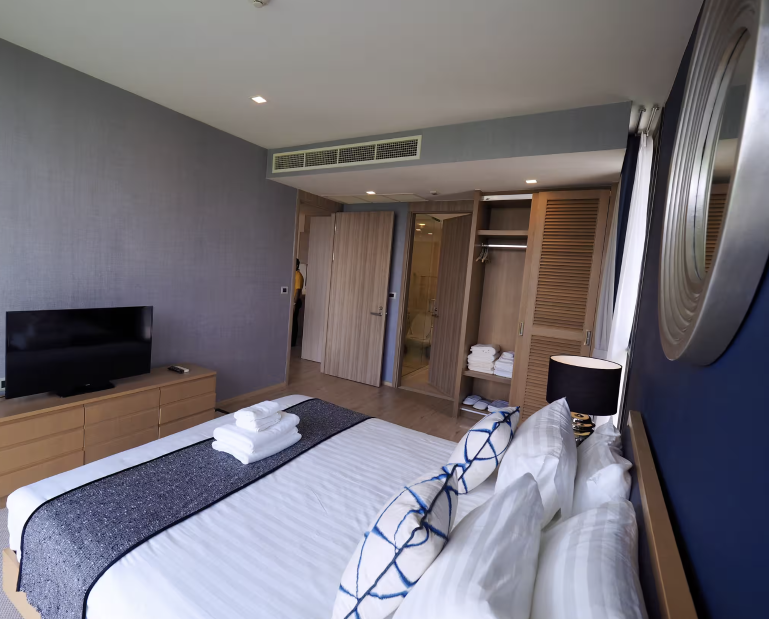 A modern hotel room with a bed, TV, and open closet with folded towels. The bed has white linens and decorative pillows, and a nightstand with a lamp. There are sliding doors leading to a bathroom or closet area, and a window with curtains.