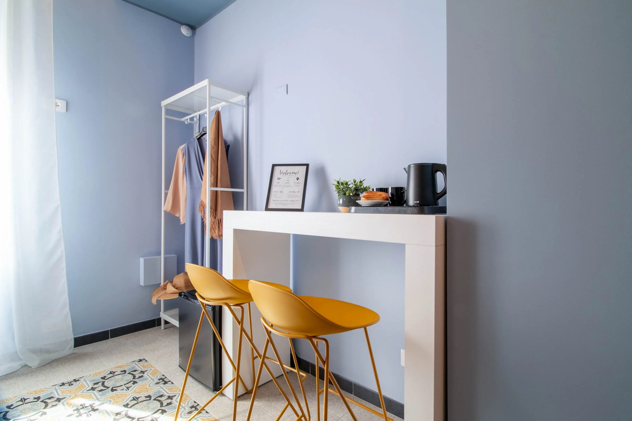Intero soggiorno con area bar, sedie gialle, cassettiera con piante e macchina per tè o caffè, quadro con scritta di benvenuto, pareti azzurre e pavimento con piastrelle decorative.