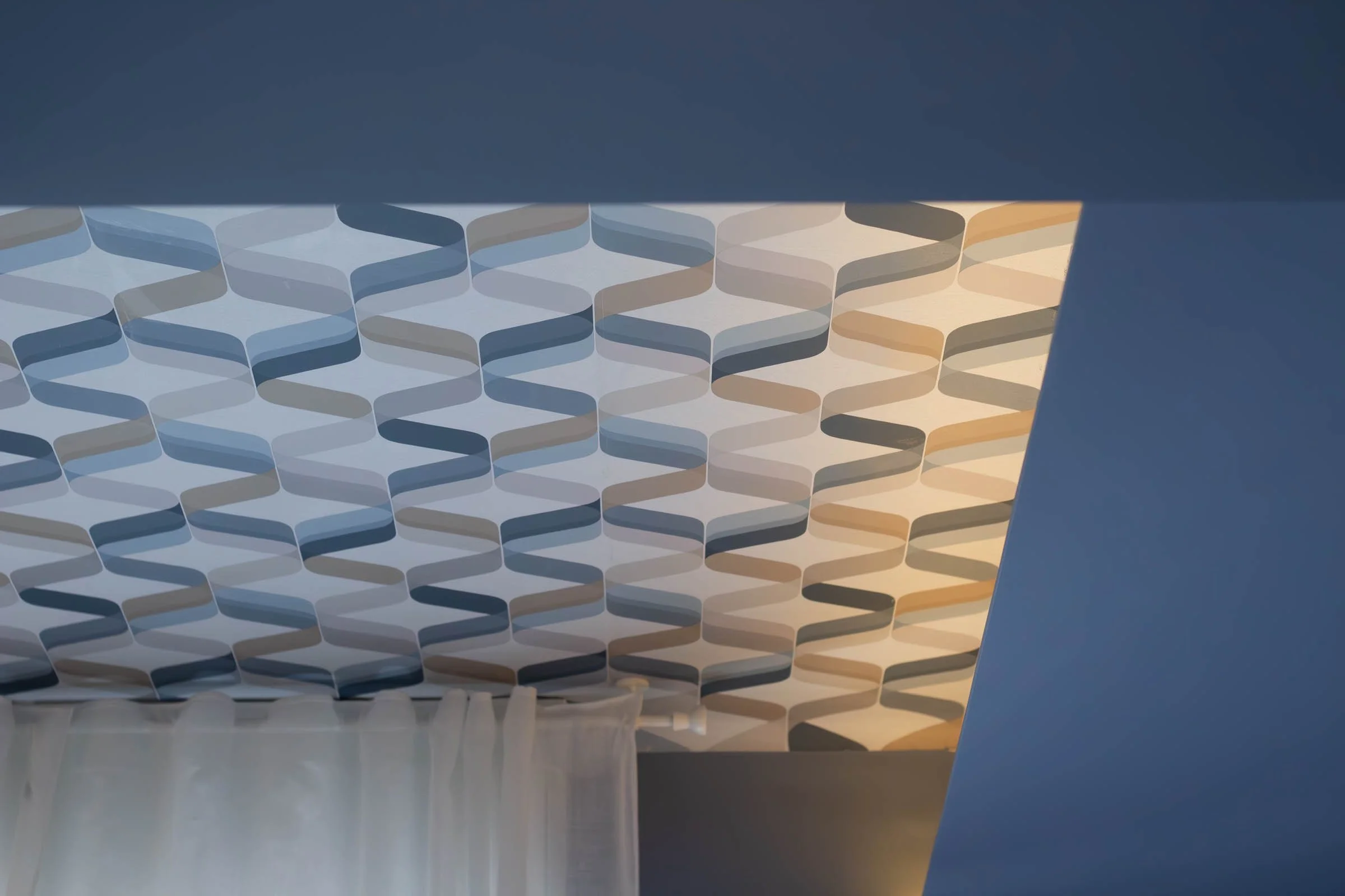 Una vista del soffitto di un edificio con un design geometrico a forma di scafo, con luci calde e tonalità di blu e beige, osservata attraverso una finestra o un’apertura.
