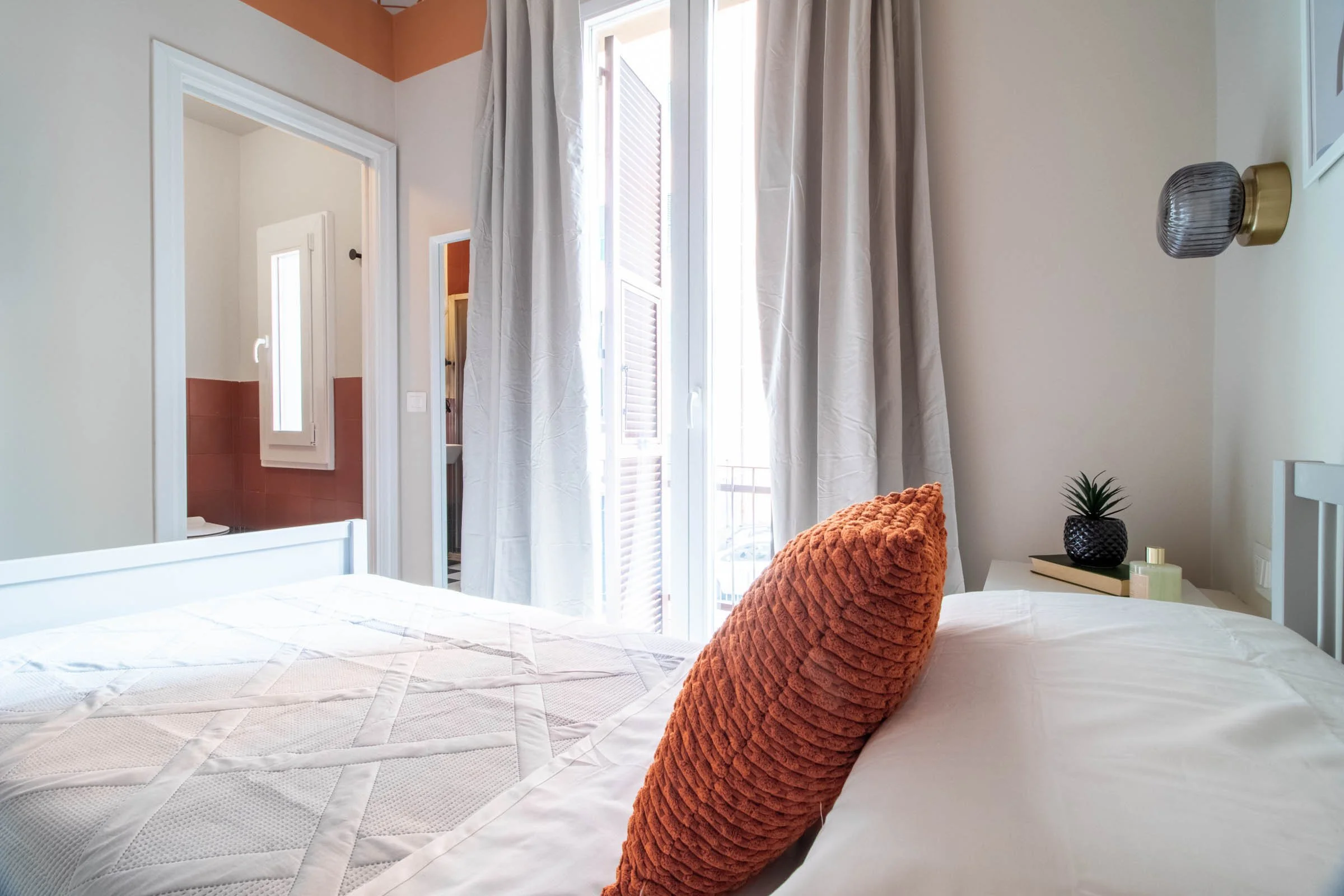 Camera da letto con grande finestra con tende bianche e vista sulla strada, letto con coperta bianca e cuscino arancione, scrivania con pianta decorativa e lampada, pareti colorate di beige e terracotta.