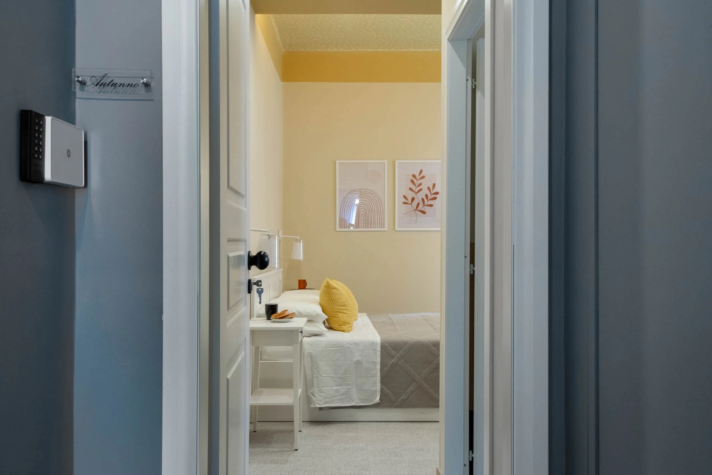 Vista di una stanza con pareti di colore giallo chiaro e decorazioni astratte, vista da un'apertura con pareti blu, mostra un letto con finiture bianche, un comodino con una tazza e alcuni accessori, e decorazioni a parete