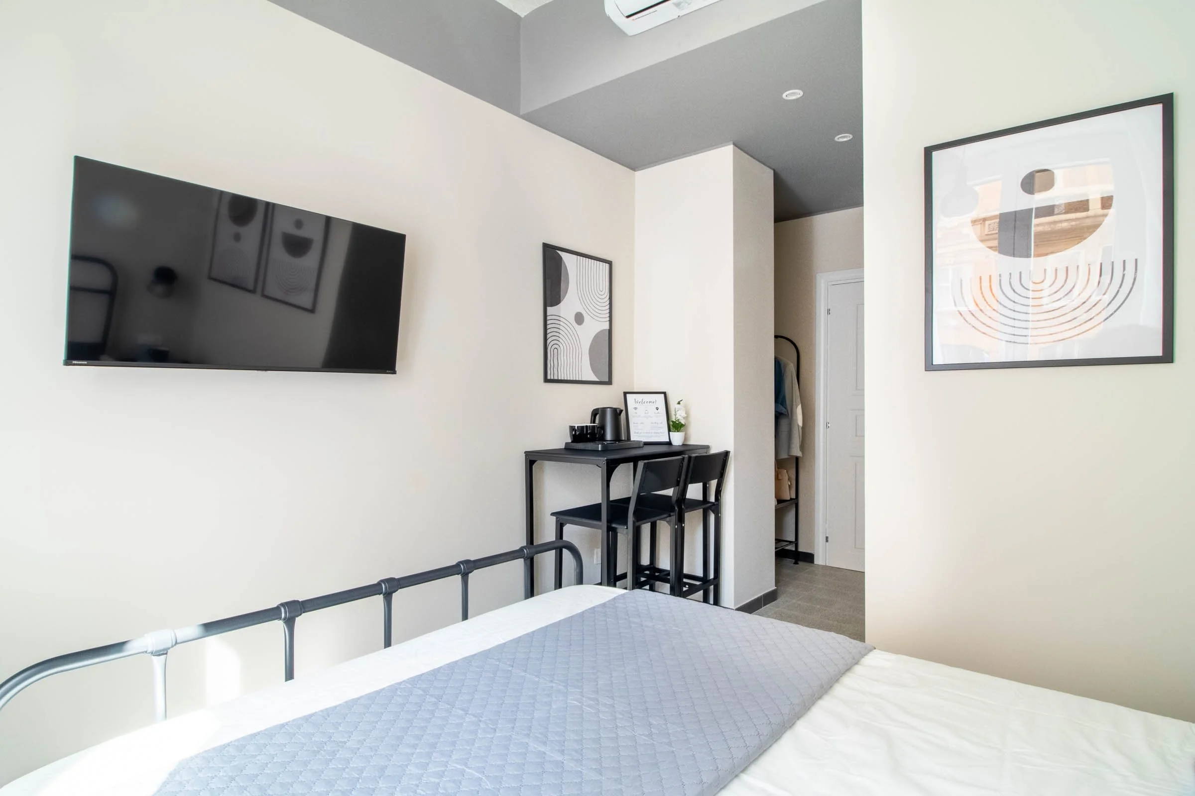 Camera da letto moderna con letto con copriletto grigio, pareti bianche, aria condizionata, opere d'arte astratte alle pareti e un tavolino con macchina da caffè e cartellina.