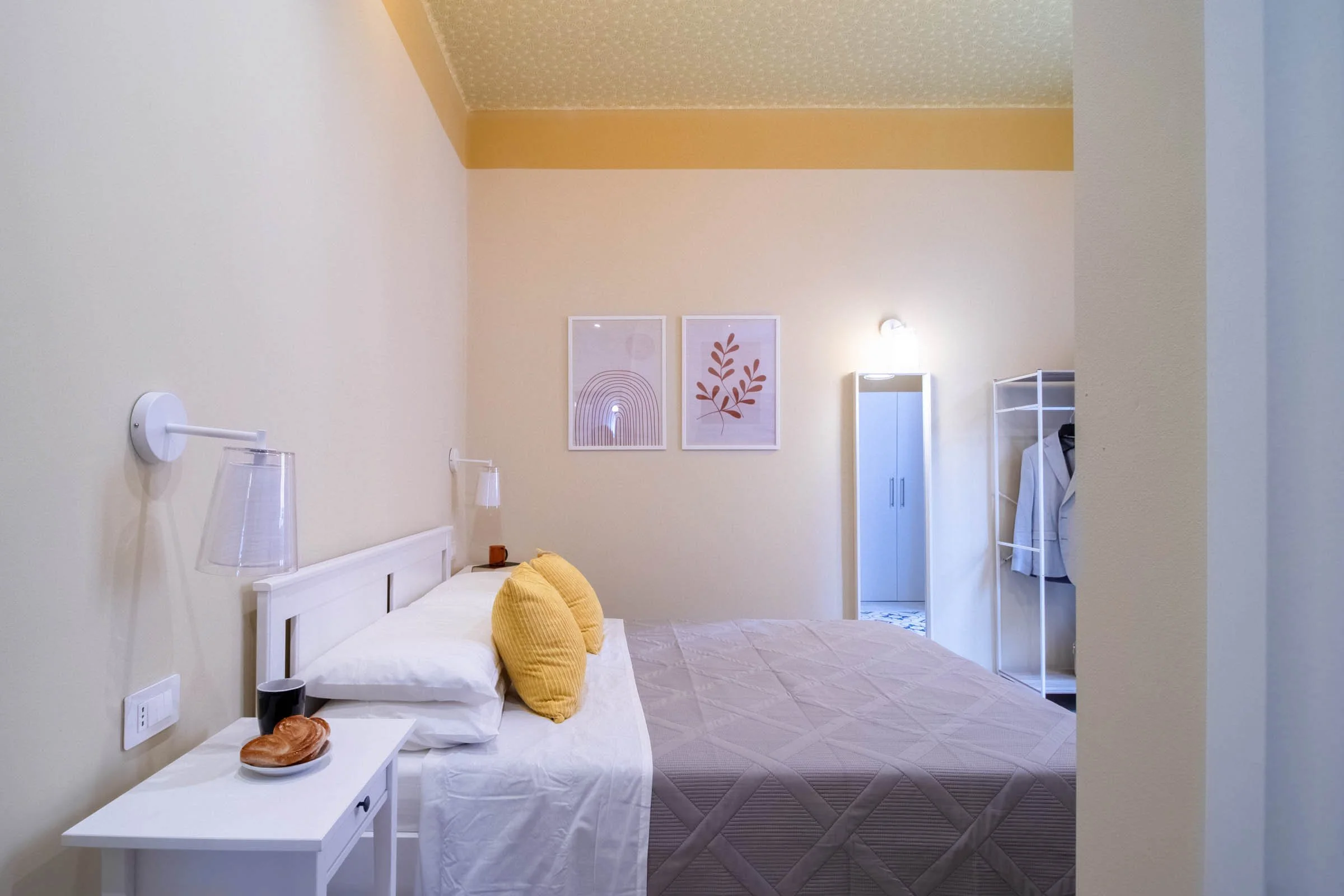 Camera da letto con letto bianco, cuscini gialli, lampade da parete, quadri con foglie stilizzate, armadio bianco e scaffale. Pareti chiare e pavimento con tappeto chiaro.