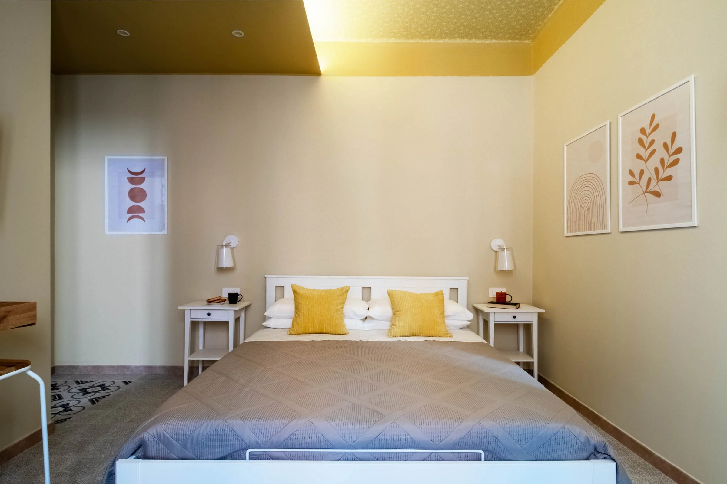 Camera da letto con letto matrimoniale, decorazioni minimaliste sulle pareti, lacune a soffitto, lampade da lettura ai lati, cuscini gialli, quadri con motivi di foglie e archi, arredamento semplice e moderno.