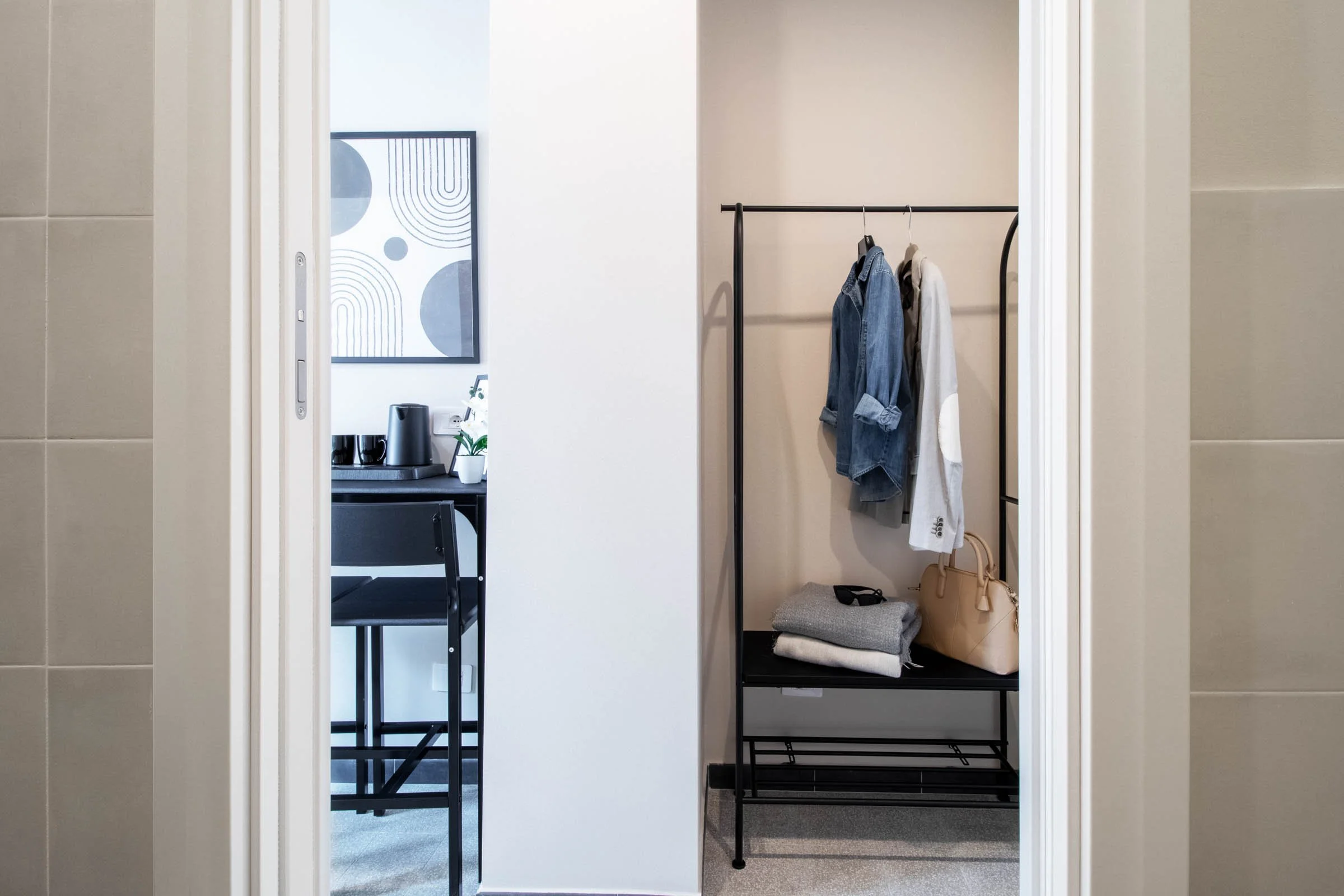 Vestibolo con appendiabiti nero che tiene una camicia di jeans e un cappotto bianco, sotto una scarpiera con un paio di scarpe nere e una borsa beige, e un facendo alla parete con tessuto grigio.