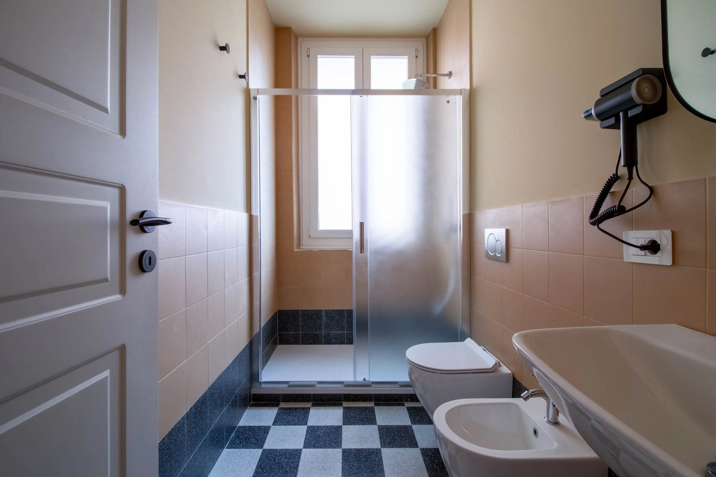 Bagno con doccia, bidet e water, finestra sulla parete opposta, asciugamani e accessori sul muro, piastrelle nere e beige.