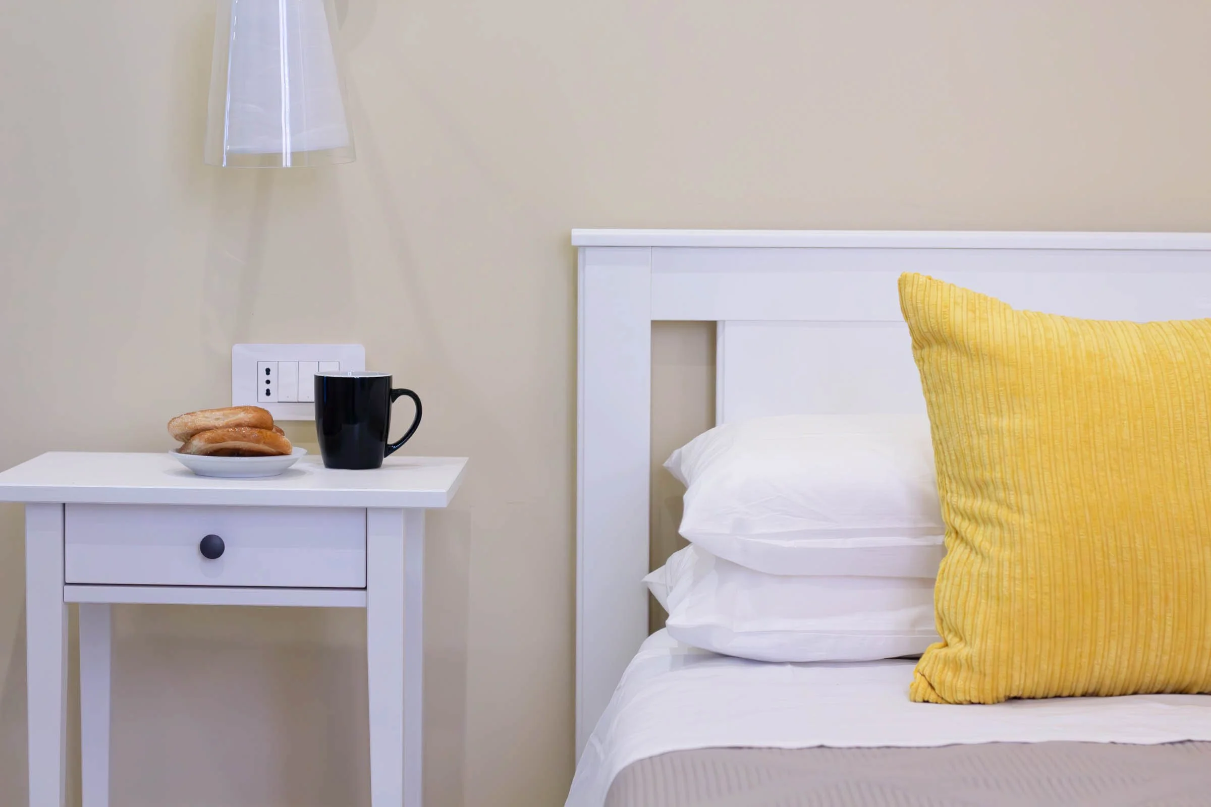 Una stanza con un letto con lenzuola bianche e un cuscino giallo a righe. Accanto al letto, una piccola scrivania bianca con una pila di ciambelle, una tazza nera e una presa elettrica sulla parete.