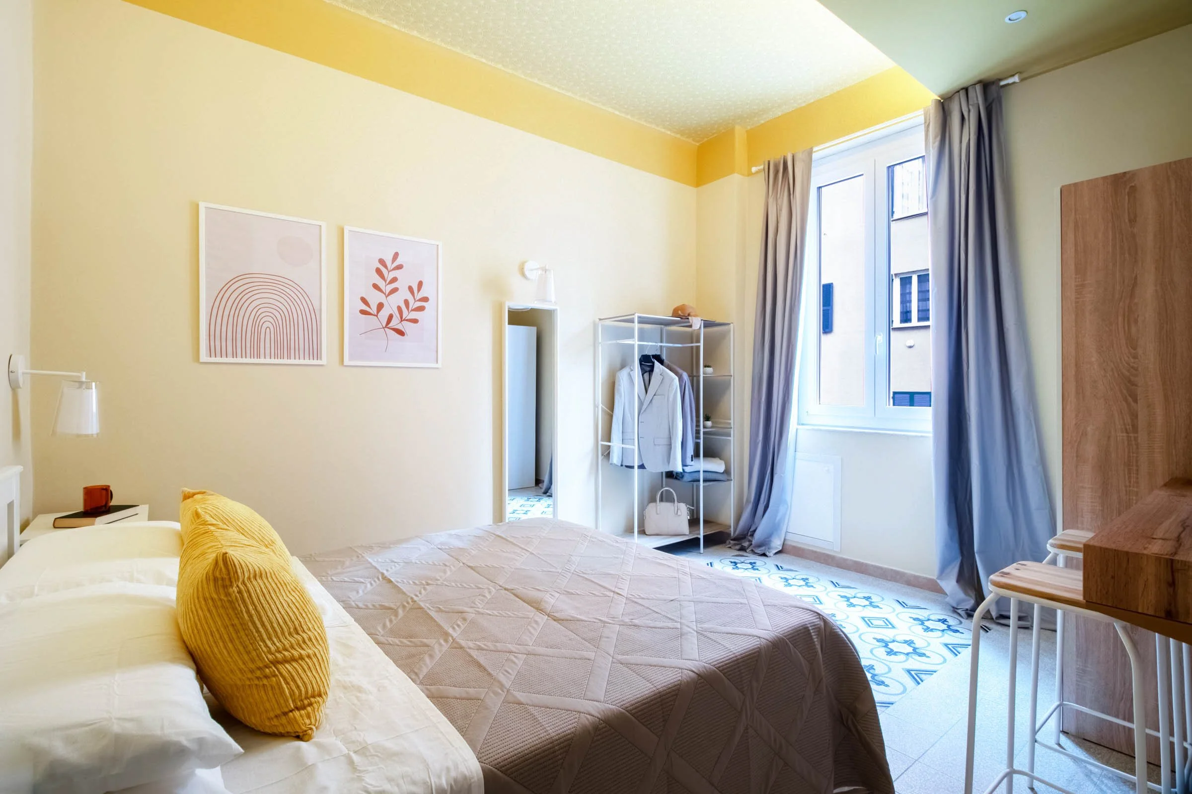 Camera da letto con letto con coperta beige e cuscini gialli, armadio aperto con vestiti, due quadri con motivi astratti e foglie, finestra con tende azzurre, arredo in stile minimalista.