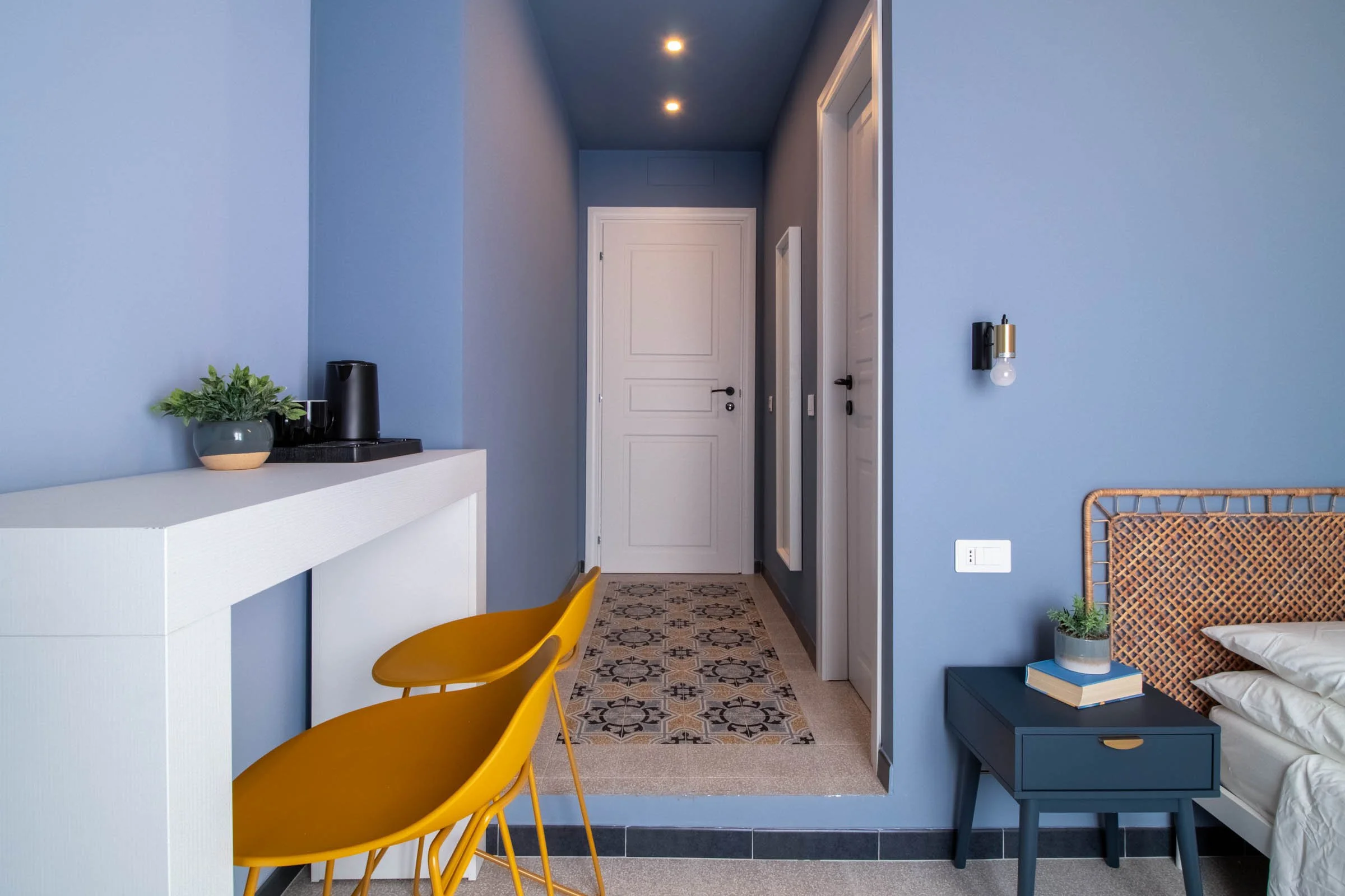 Interno di una stanza con pareti azzurre, porta bianca in fondo e pavimento con piastrelle decorative. In primo piano, due sedie gialle, una piccola consolle bianca con pianta e macchina da caffè, e a destra una panca blu con un libro e una pianta, a
