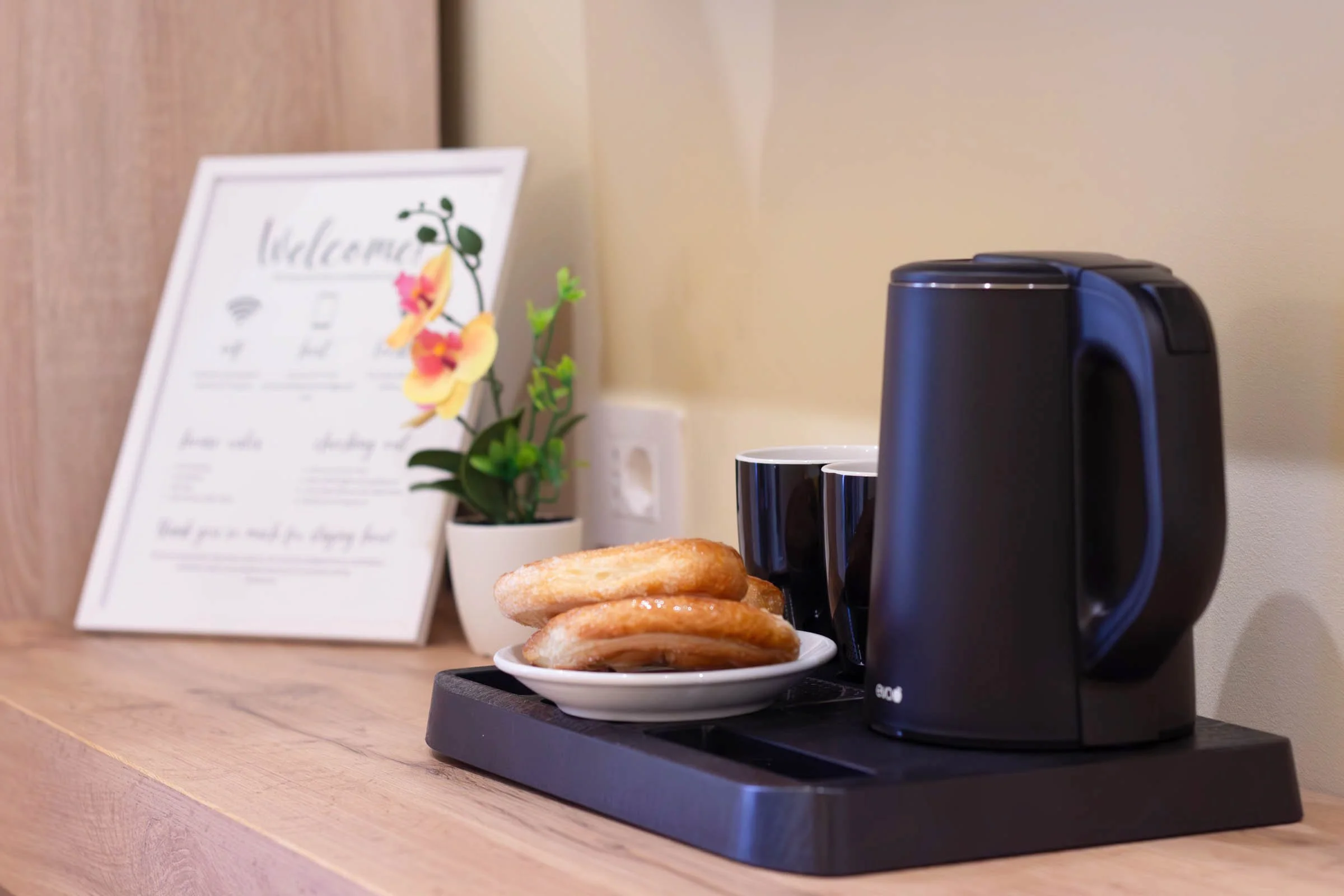 Macchina per il caffè con tazzine, ciambelle su un piatto, e un cartello con menu dietro, su una superficie in legno, parete beige sullo sfondo.