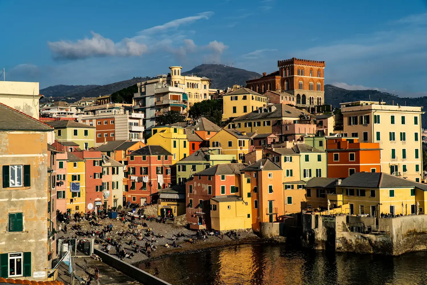 Paesaggio urbano con case colorate su una collina e un mare in primo piano, con montagne sullo sfondo.