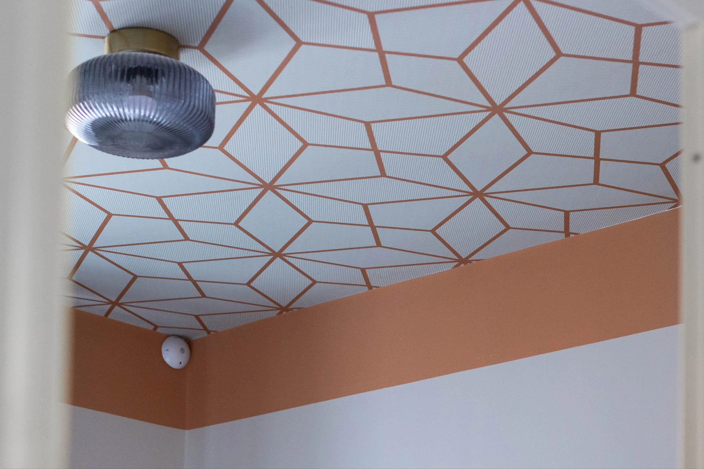 L'immagine mostra un soffitto con un motivo geometrico a rombi e linee sottili di colore rosso. Sul lato sinistro si vede una lampada cilindrica dallo stile moderno e di colore beige. In basso a sinistra, c'è uno speaker bianco, e sul pavimento di co